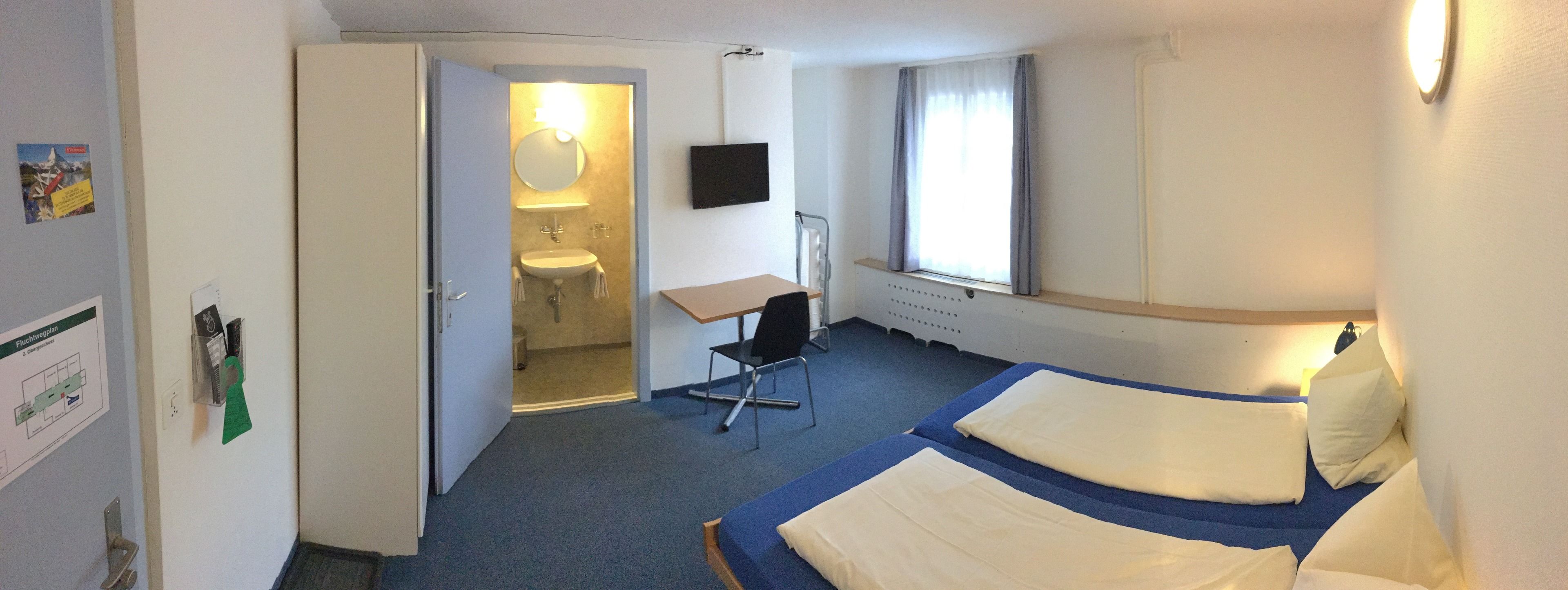 Habitación de hotel con dos camas, un escritorio, una silla, un lavabo y un televisor.