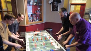 A group of people playing foosball in a room with a red and white color scheme, a glass display case with various items, and a sign indicating free booking of hostels at reception.