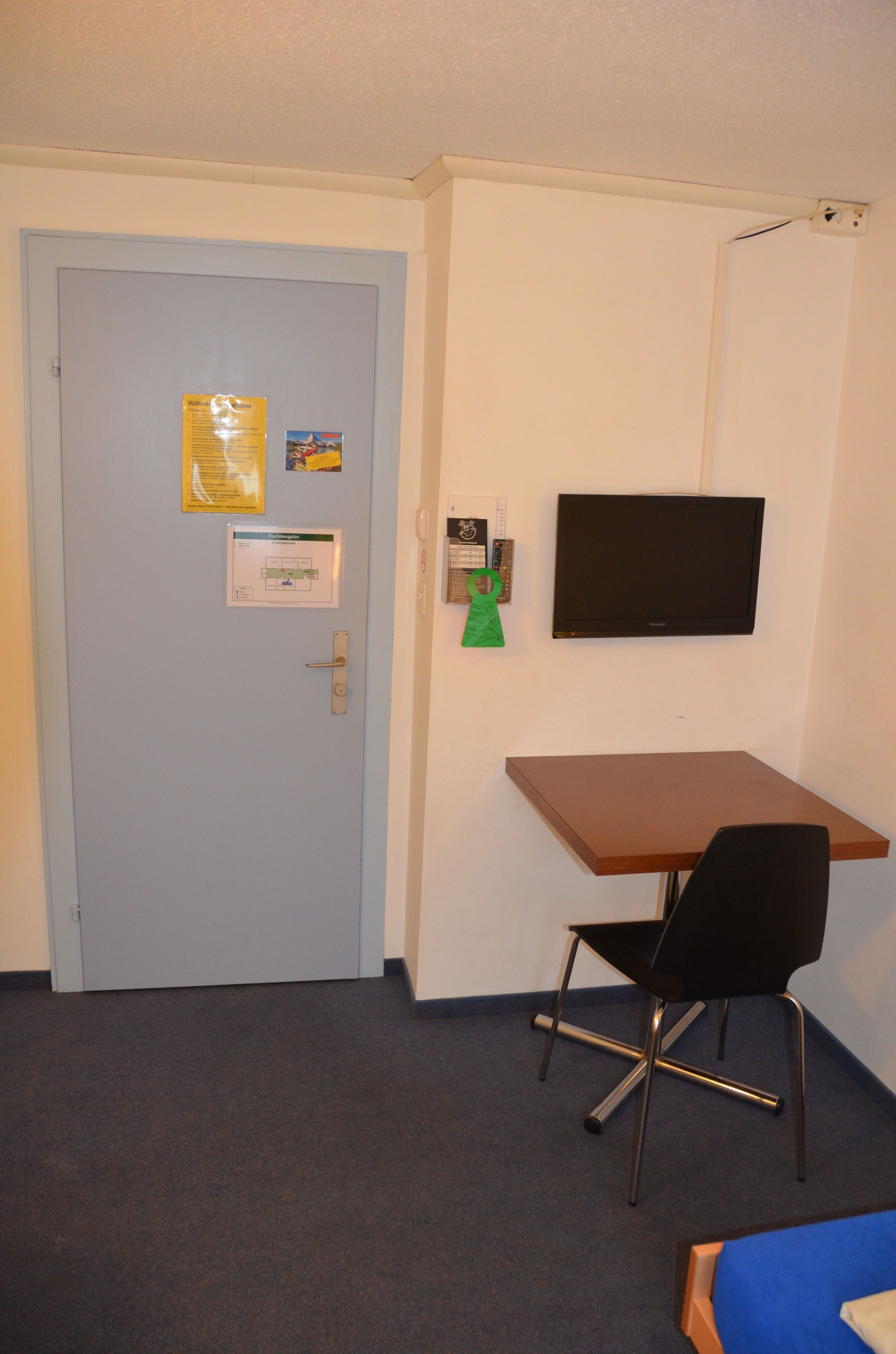 A room with a light blue door, a desk with a black chair, a television mounted on the wall, and a bed with a blue cover.