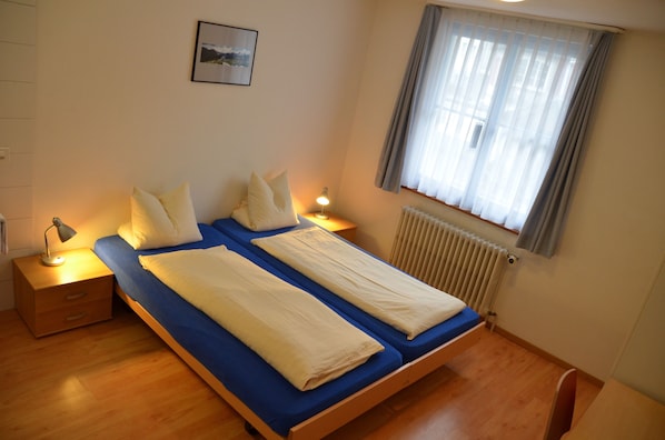 A double bed with a blue mattress and white linens, two bedside tables with lamps, a window with curtains, and a radiator. - Hirschen Schwyz GmbH - Hostel (Schwyz)