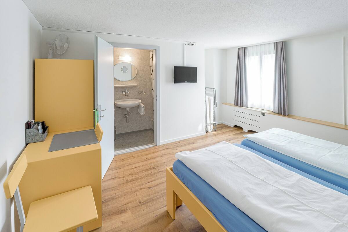 A hotel room with a bed, a desk, a chair, a bathroom with a sink and mirror, and a TV on the wall.