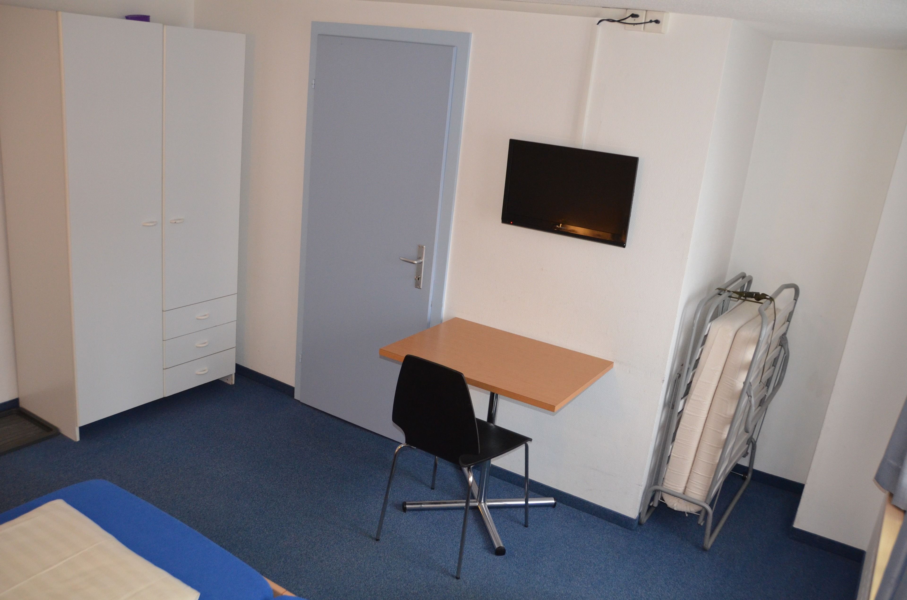A small room with a single bed, a desk, a chair, a wardrobe, and a TV mounted on the wall.
