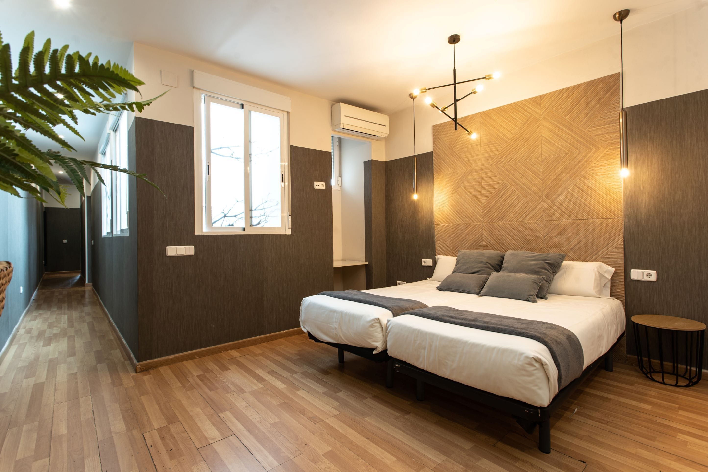 A modern bedroom with a wooden floor, two beds, a large window, and a ceiling light fixture.