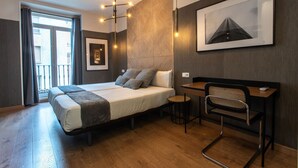 A modern hotel room with a large bed, a desk, and a chair. There is a window with curtains, a wall-mounted picture, and a ceiling light fixture. - Suites You Zinc (Madrid)
