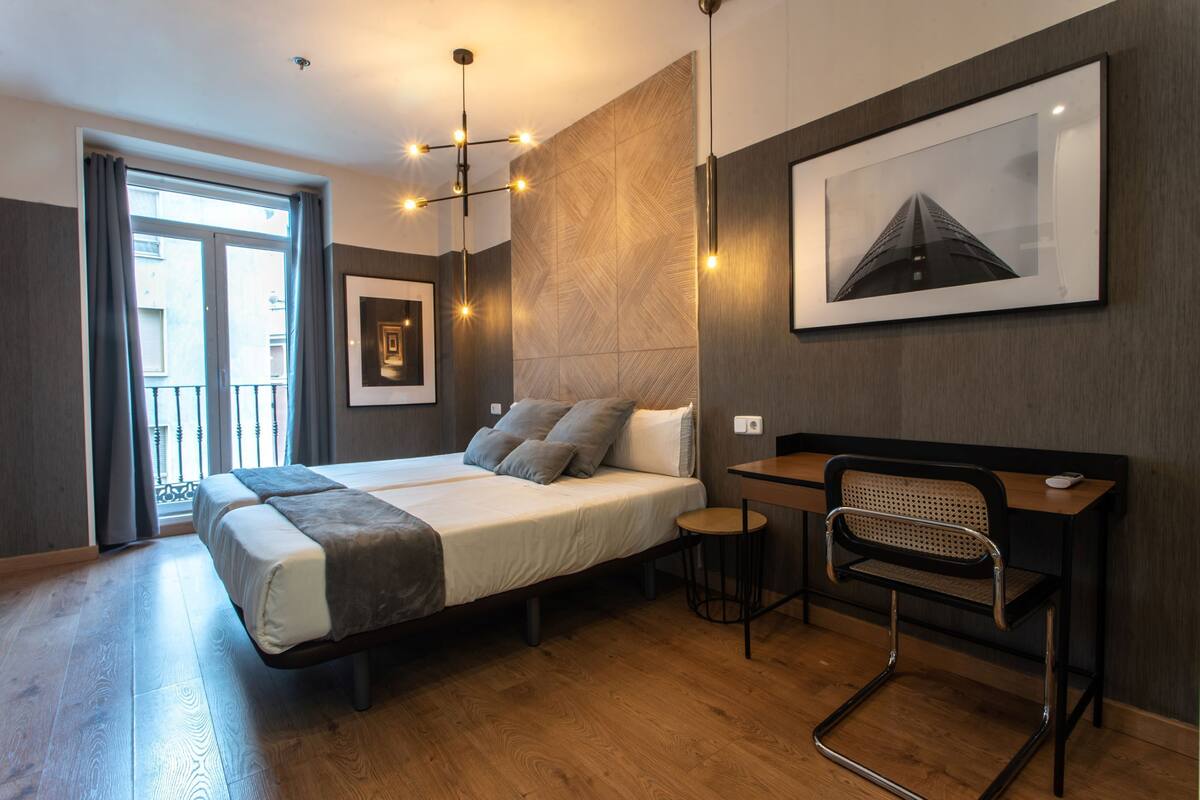 A modern hotel room with a large bed, a desk, and a chair. There is a window with curtains, a wall-mounted picture, and a ceiling light fixture.