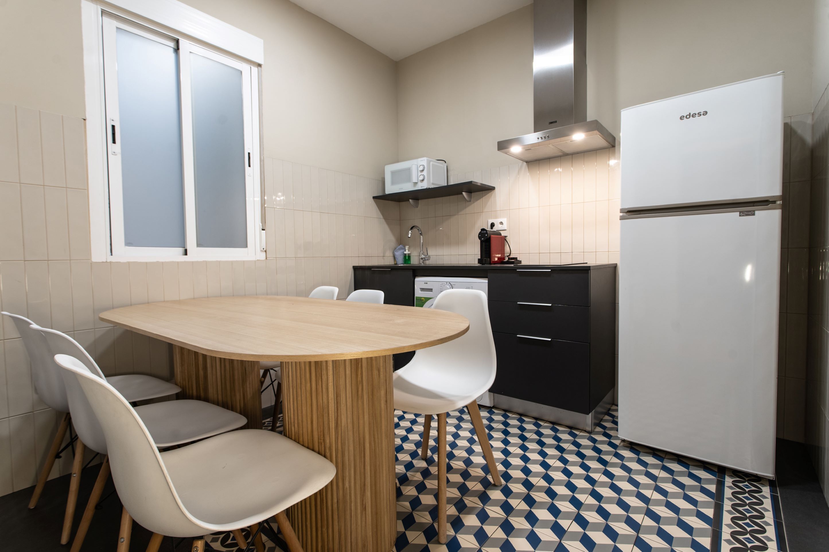 A modern kitchen with a round wooden table, white chairs, a microwave, a dishwasher, and a refrigerator.