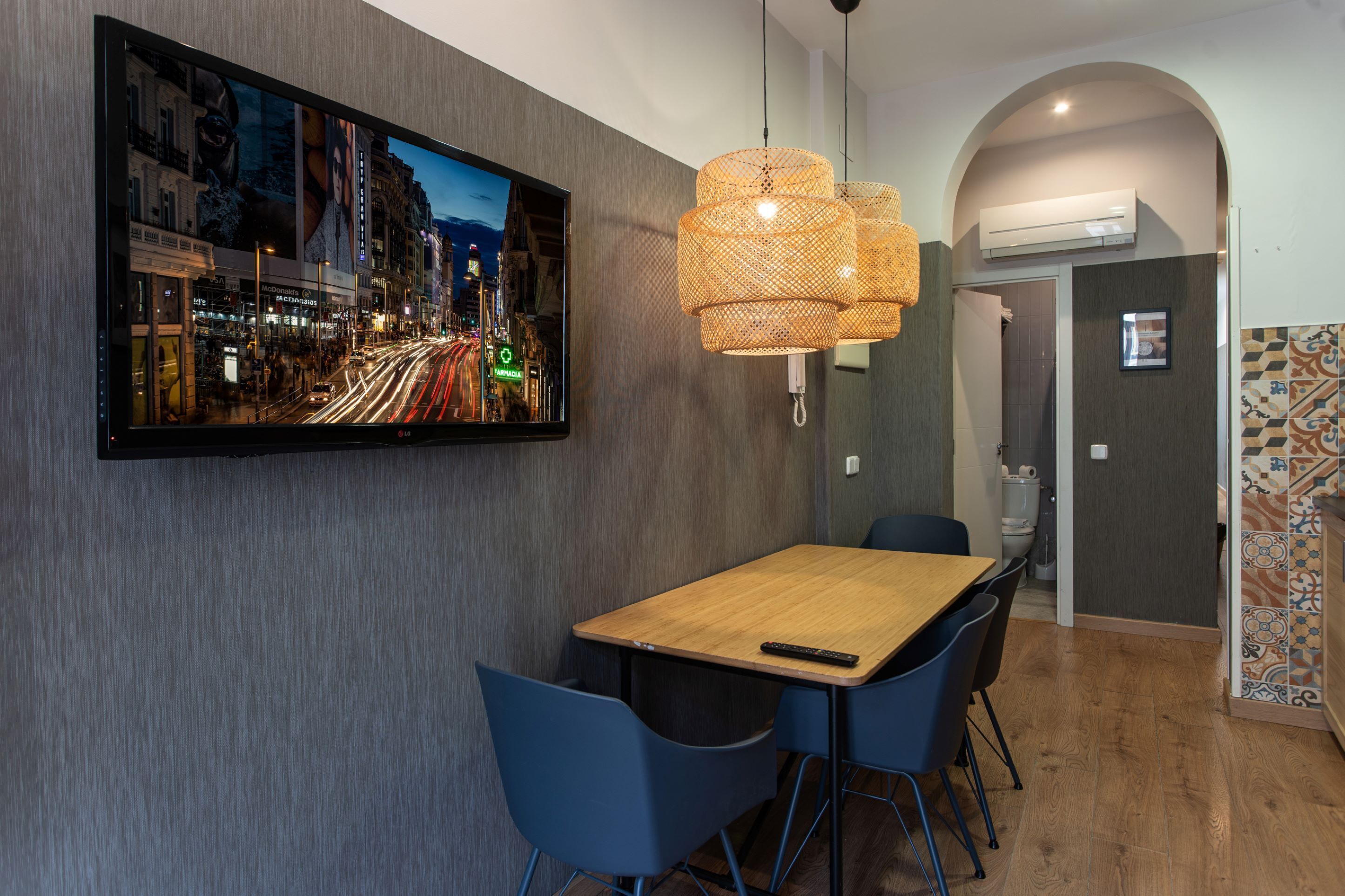 A modern dining area with a wooden table, blue chairs, a large flat-screen TV on the wall, and a woven pendant light.