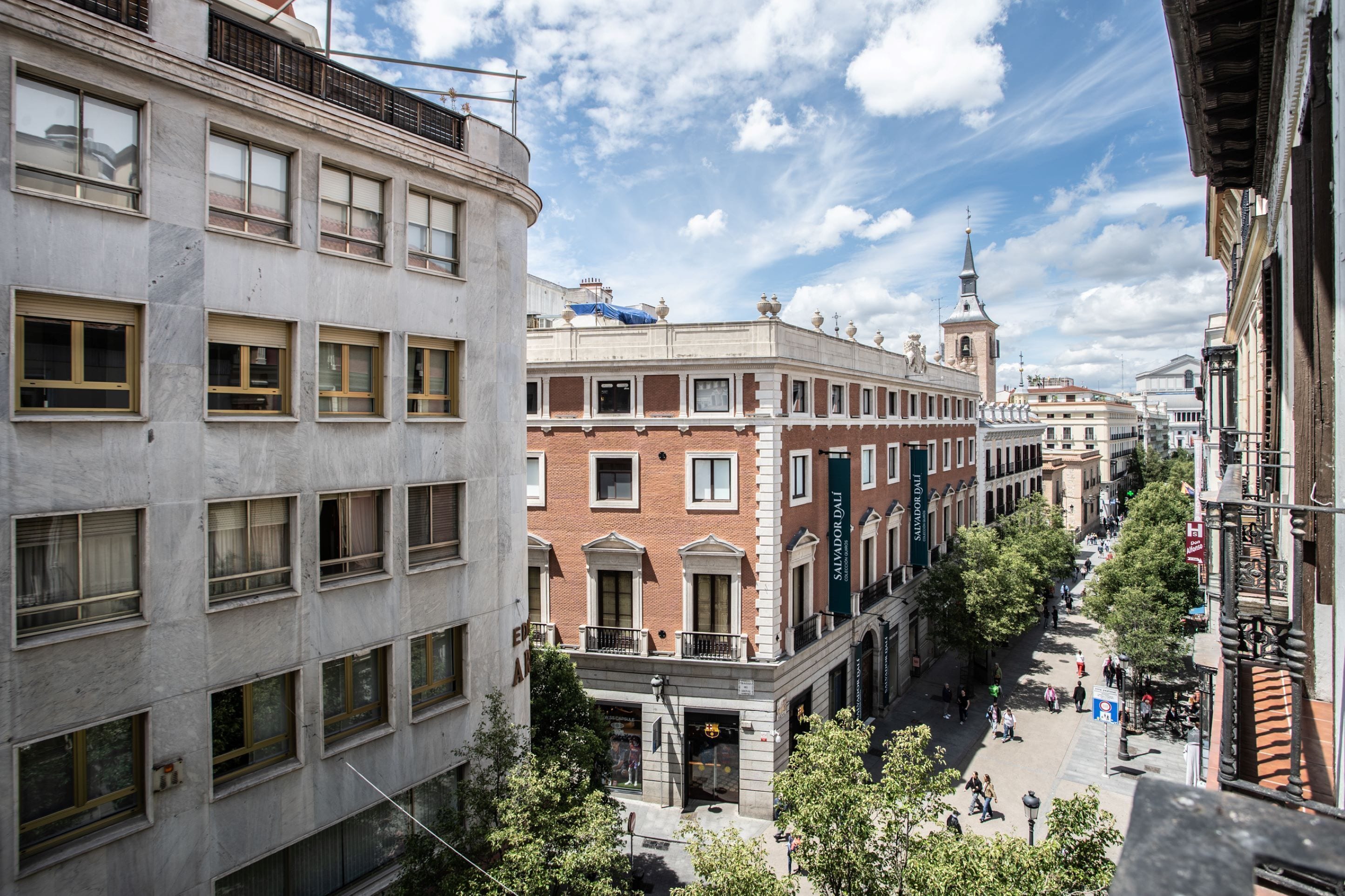 A street view with a mix of modern and historic buildings, a prominent church tower, and a pedestrian area with trees and people.