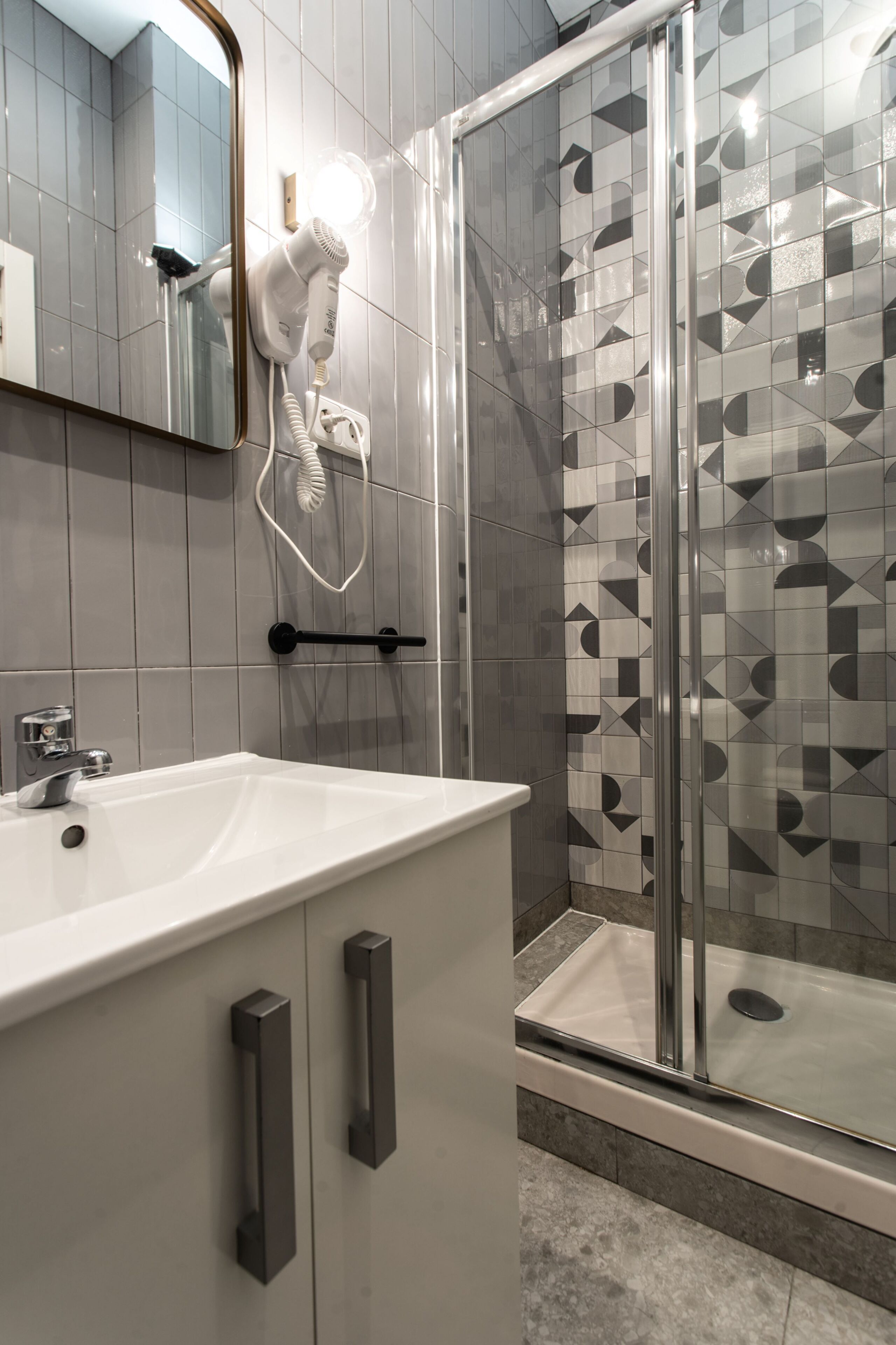 A modern bathroom with a white sink, a hairdryer, and a shower with geometric-patterned tiles.