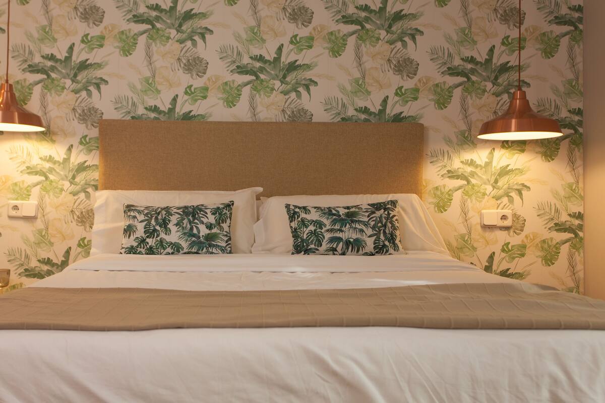 A bed with a headboard, two pillows with a tropical leaf pattern, and two pendant lights.
