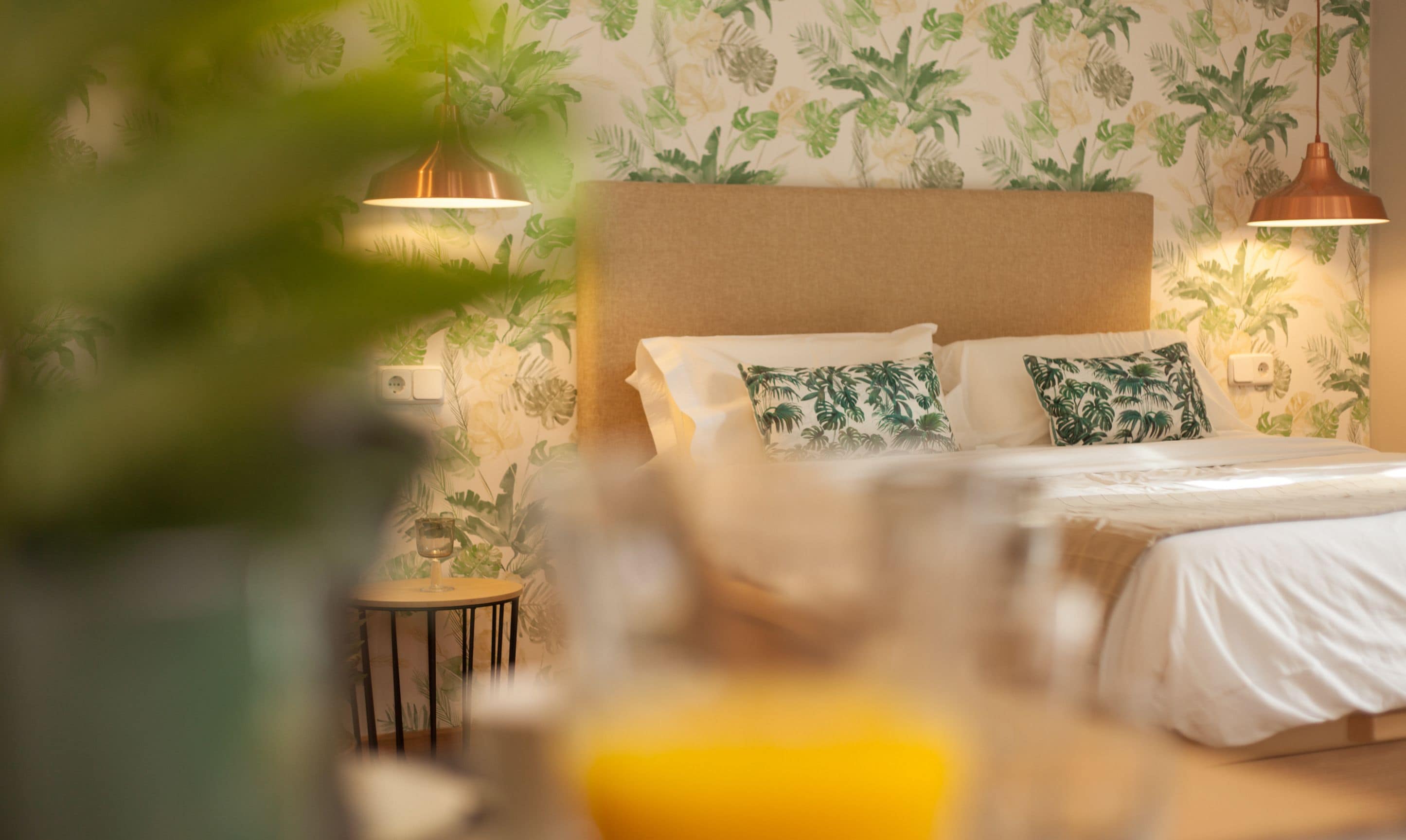 A hotel room with a bed, two wall-mounted lamps, a small round table, and a potted plant.