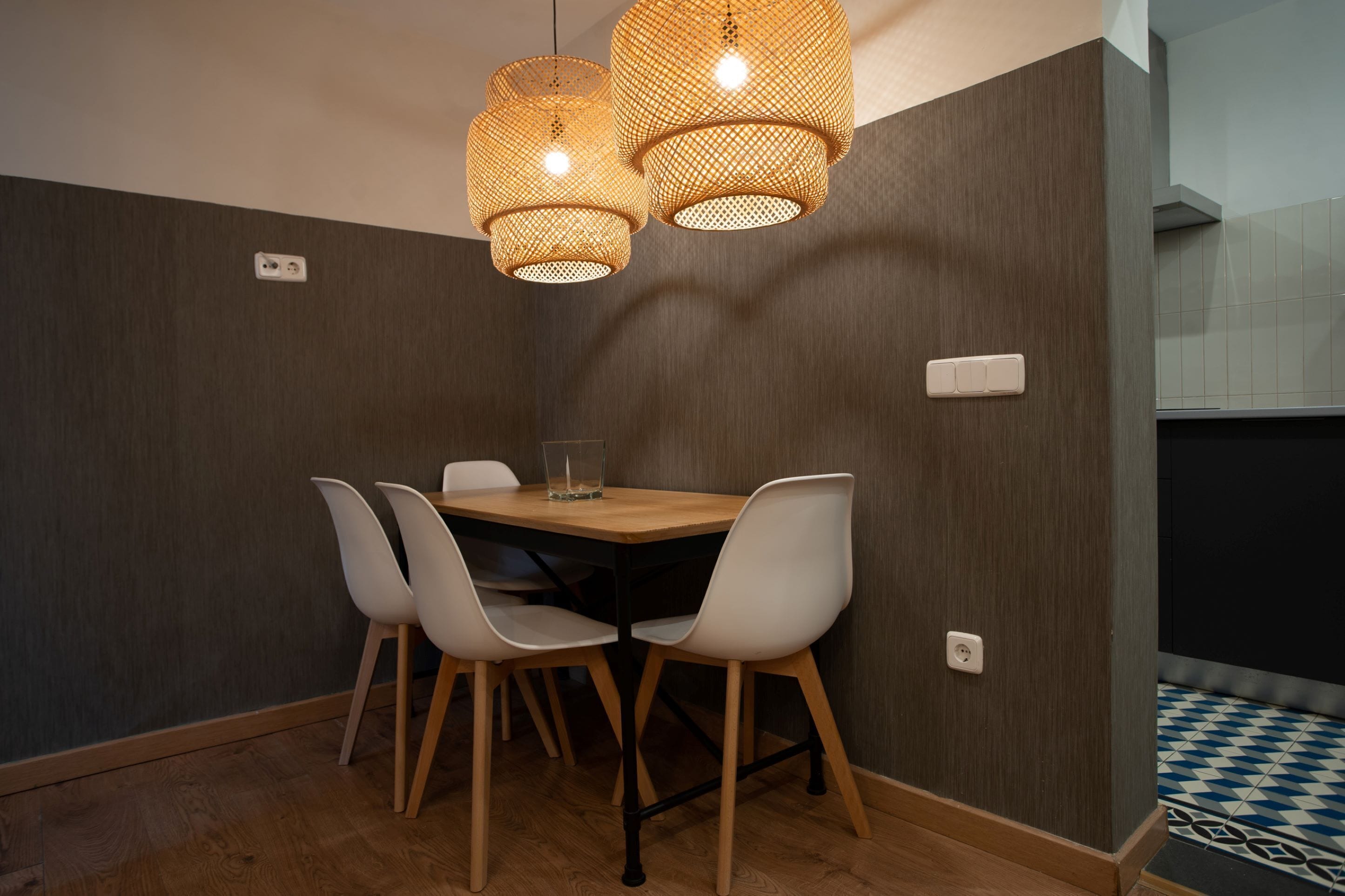 Un comedor con mesa de madera y cuatro sillas, dos lámparas colgantes y piso de baldosas.