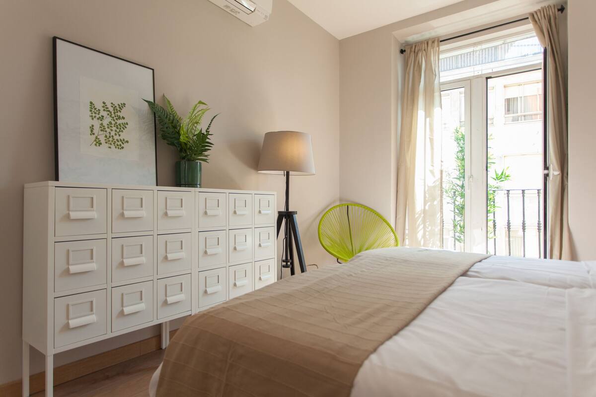 A bedroom with a bed, a white dresser, a lamp, a green chair, and a window with curtains.