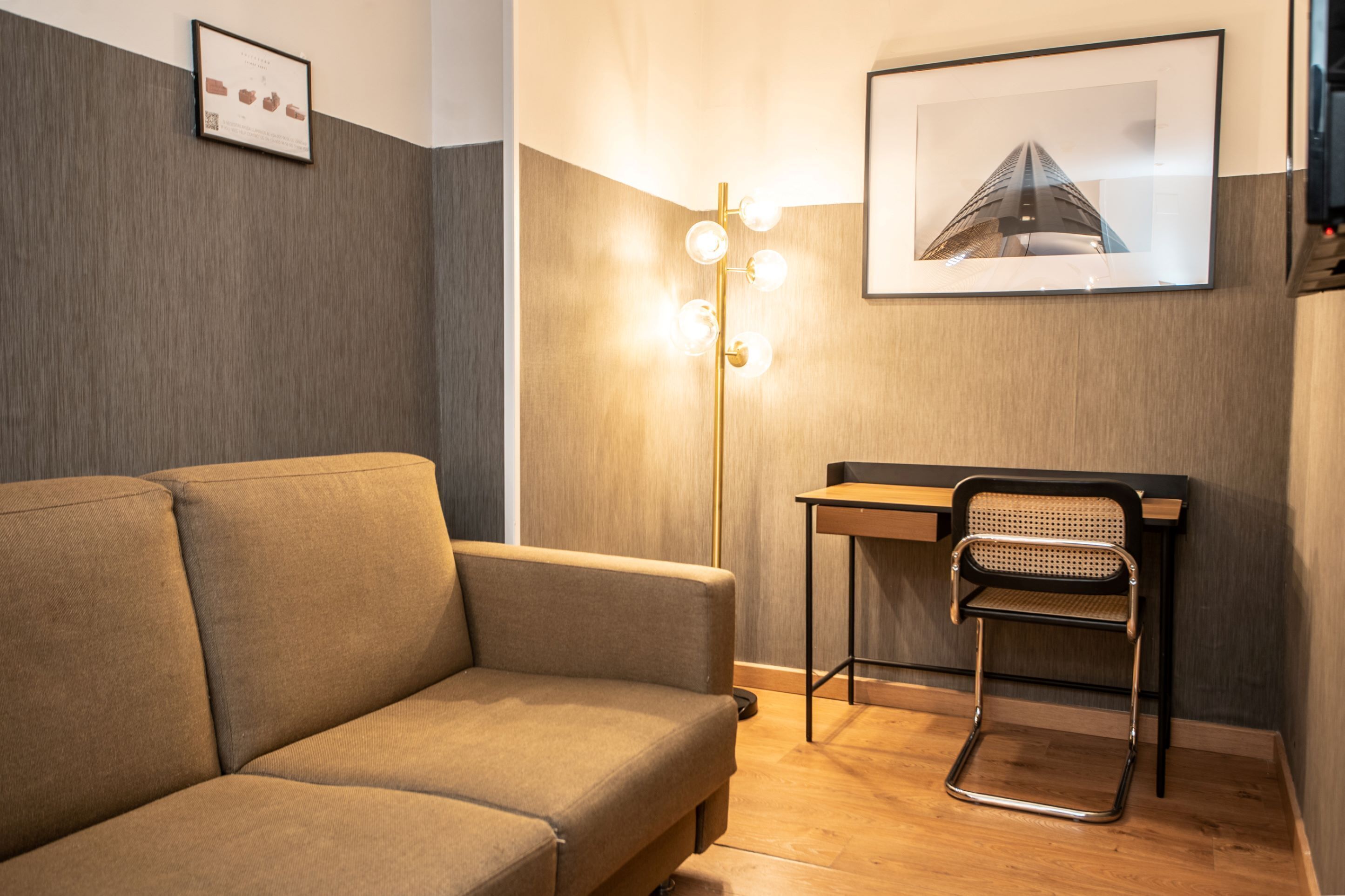Ein modernes Zimmer mit einer Couch, einem Schreibtisch und einem Sessel. An der Wand hängt ein gerahmtes Bild und eine Stehlampe mit mehreren Leuchten.