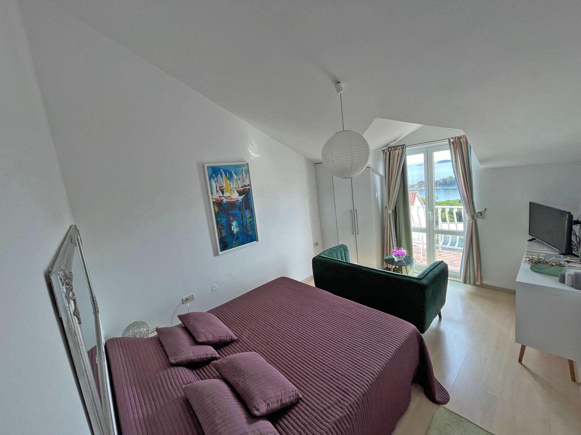 A bedroom with a bed, a desk, a chair, a TV, a painting, and a view of the outside.