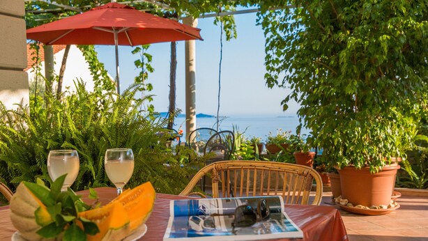 Un espace de restauration en plein air avec une table dressée pour deux personnes, un parasol rouge et une vue sur la mer.