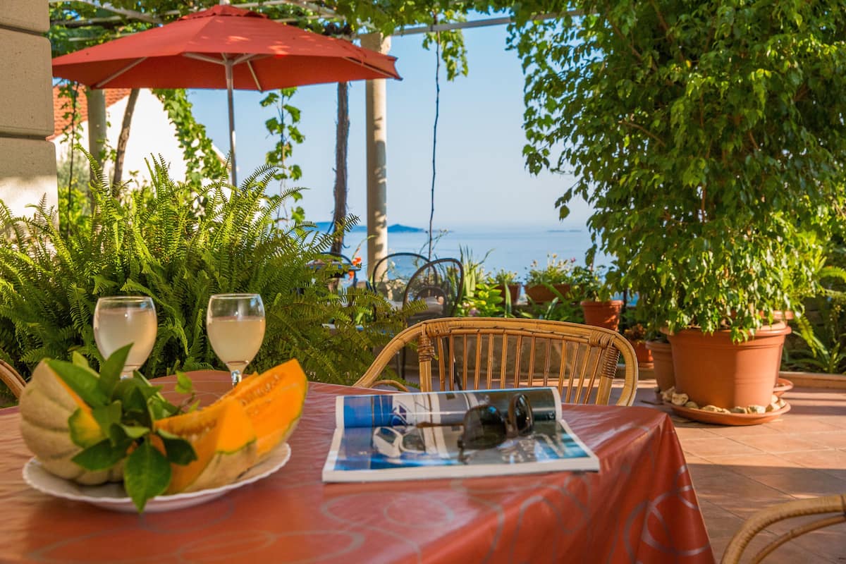 Un área de comedor al aire libre con una mesa para dos, una sombrilla roja y vista al mar.
