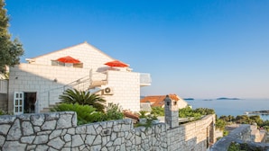 Un bâtiment blanc avec un mur de pierre à l’avant, un balcon orné de parasols rouges et une vue sur la mer.