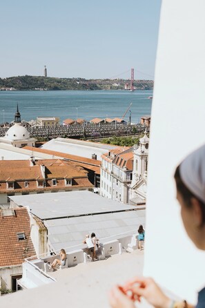 A view of a coastal cityscape with a prominent bridge in the background, a dome-shaped building, and a person sitting on a rooftop. - Lisb'on Hostel (Lisbon)