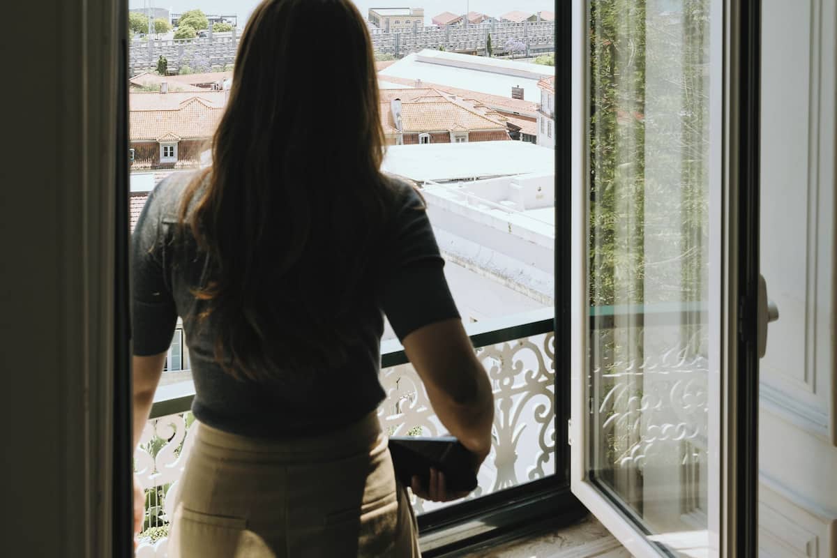 Una persona de pie junto a una ventana con vista a un paisaje urbano que incluye edificios y una masa de agua.