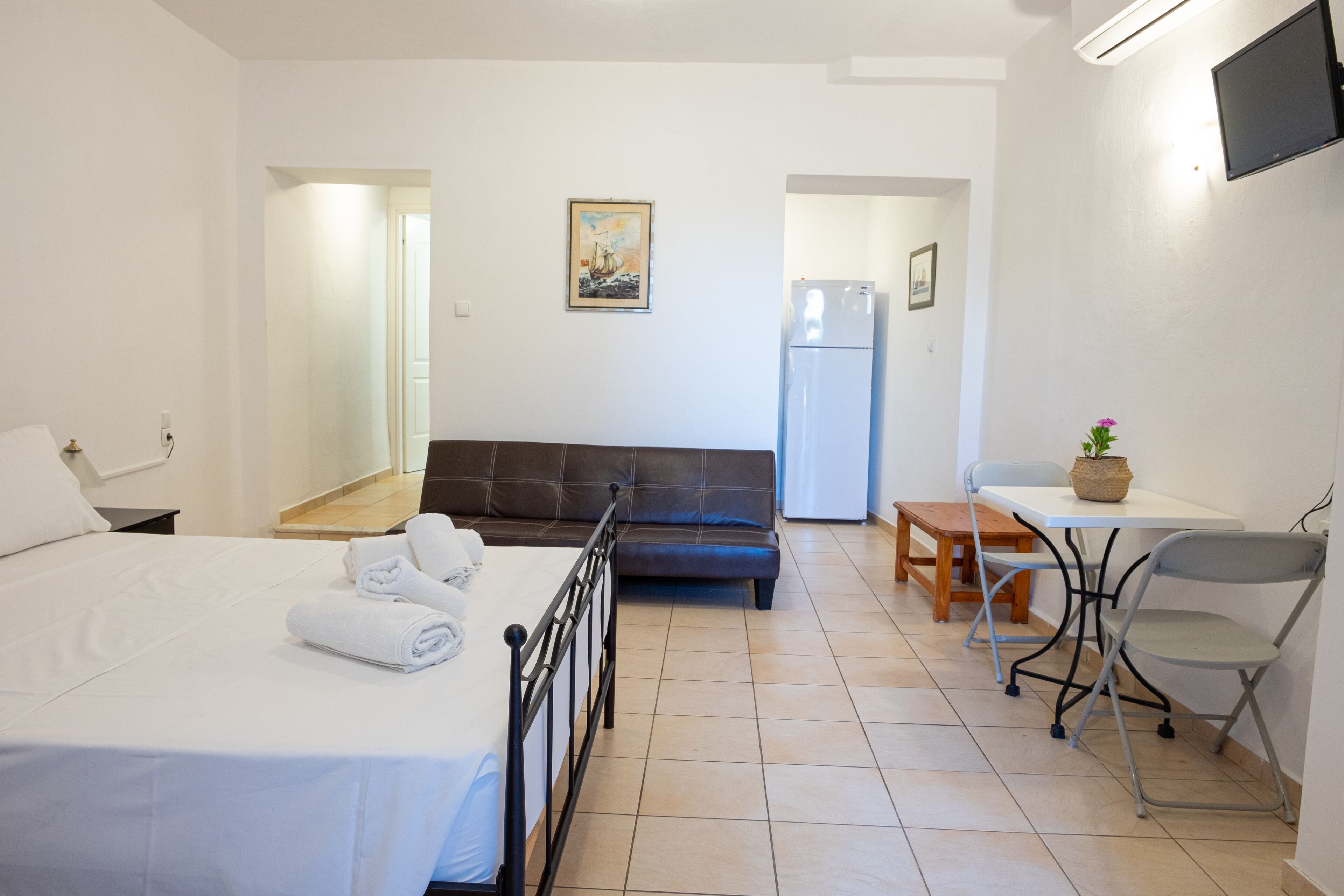 A hotel room with a bed, a sofa, a dining table with chairs, a small table with a plant, and a refrigerator.
