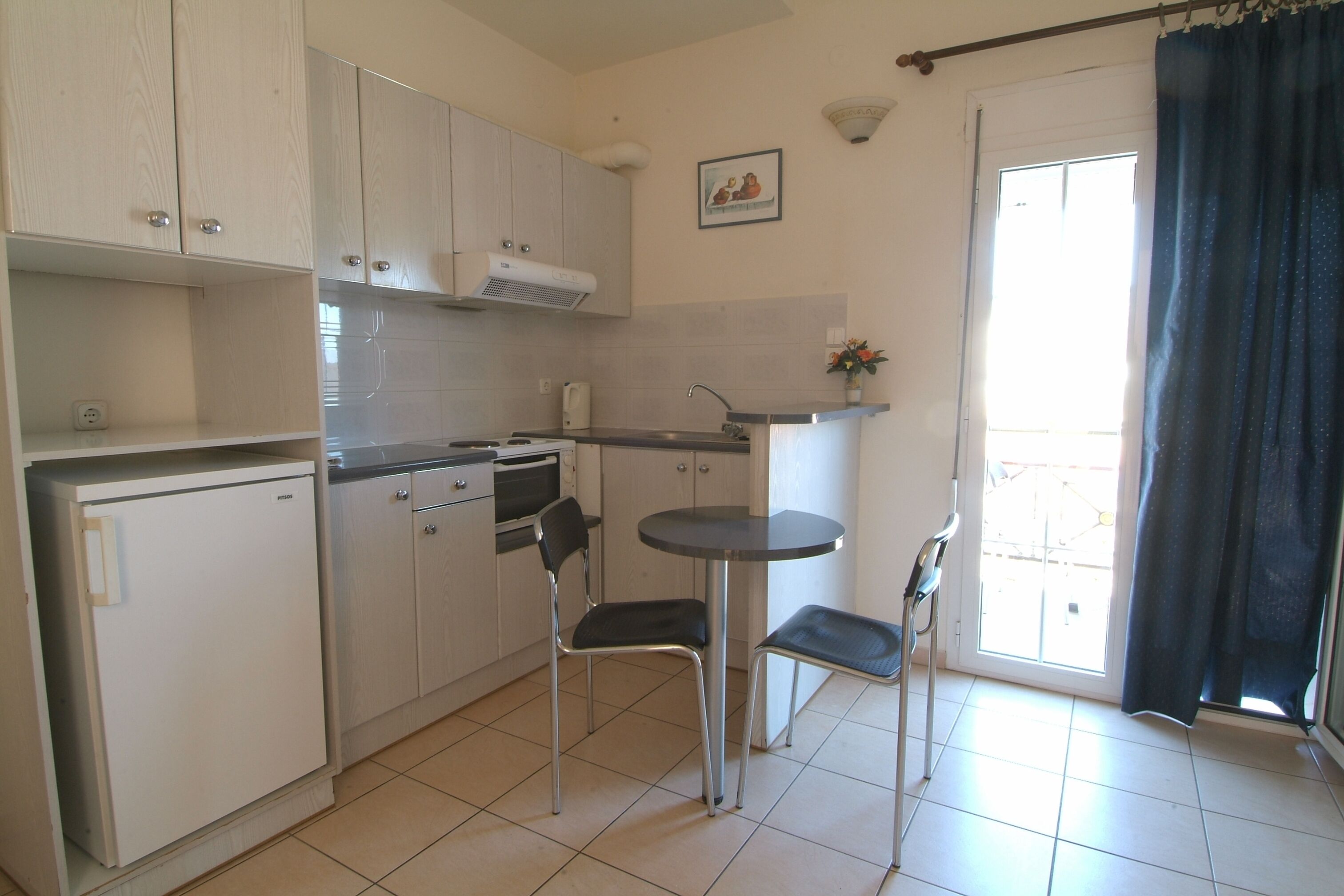 A compact kitchen with white cabinets, a small refrigerator, a sink, and a dining area with two chairs and a small table.