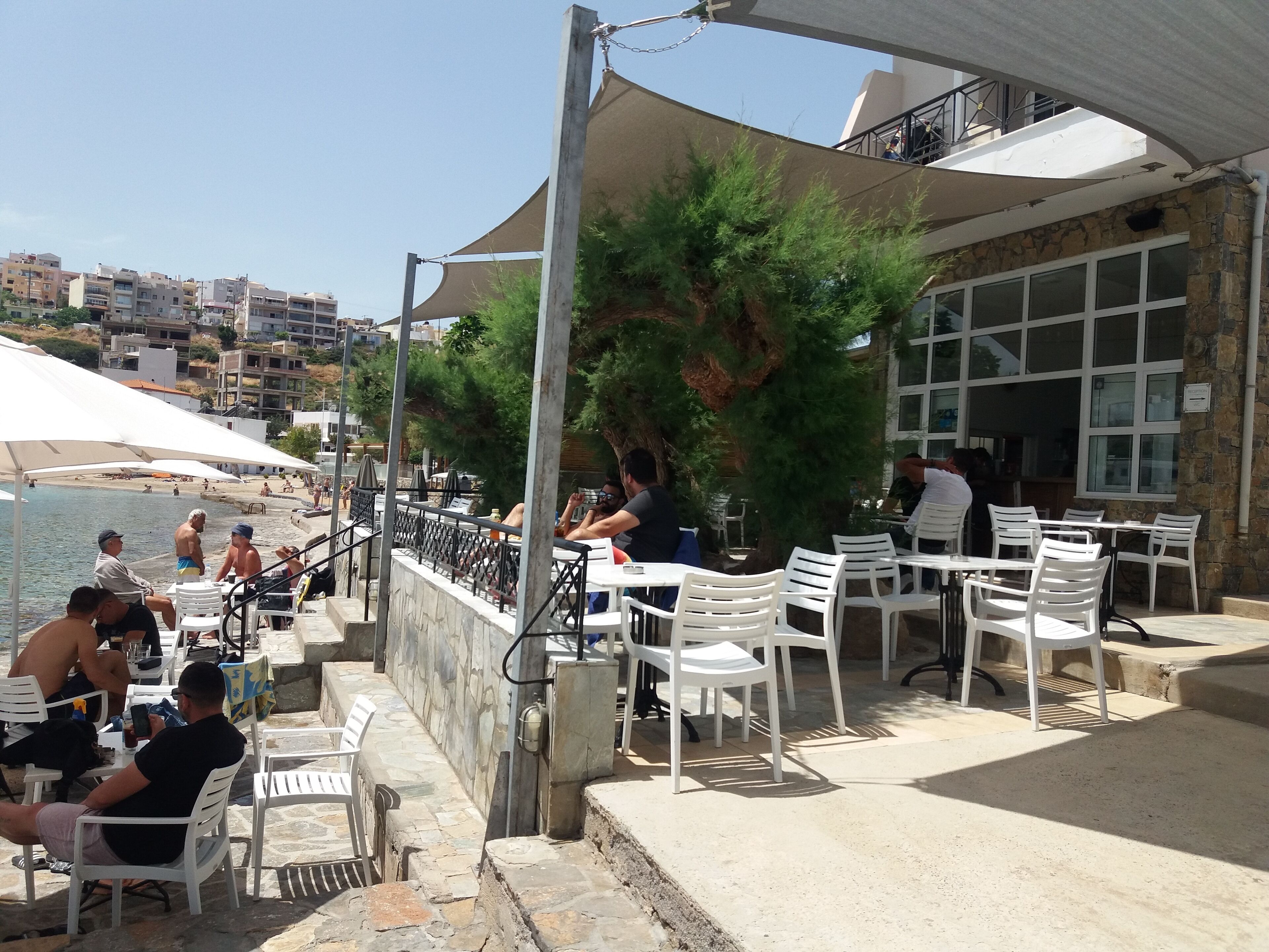 An outdoor seating area with white plastic chairs and tables, overlooking a beach and a cityscape.
