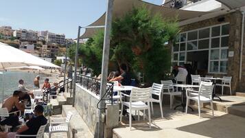 An outdoor seating area with white plastic chairs and tables, overlooking a beach and a cityscape.