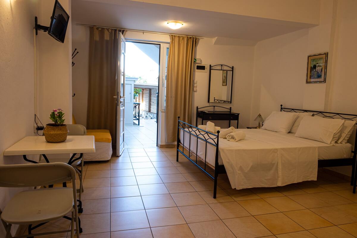 Habitación de hotel con cama, escritorio con silla, una mesita con planta, televisor montado en la pared y ventana con cortinas.
