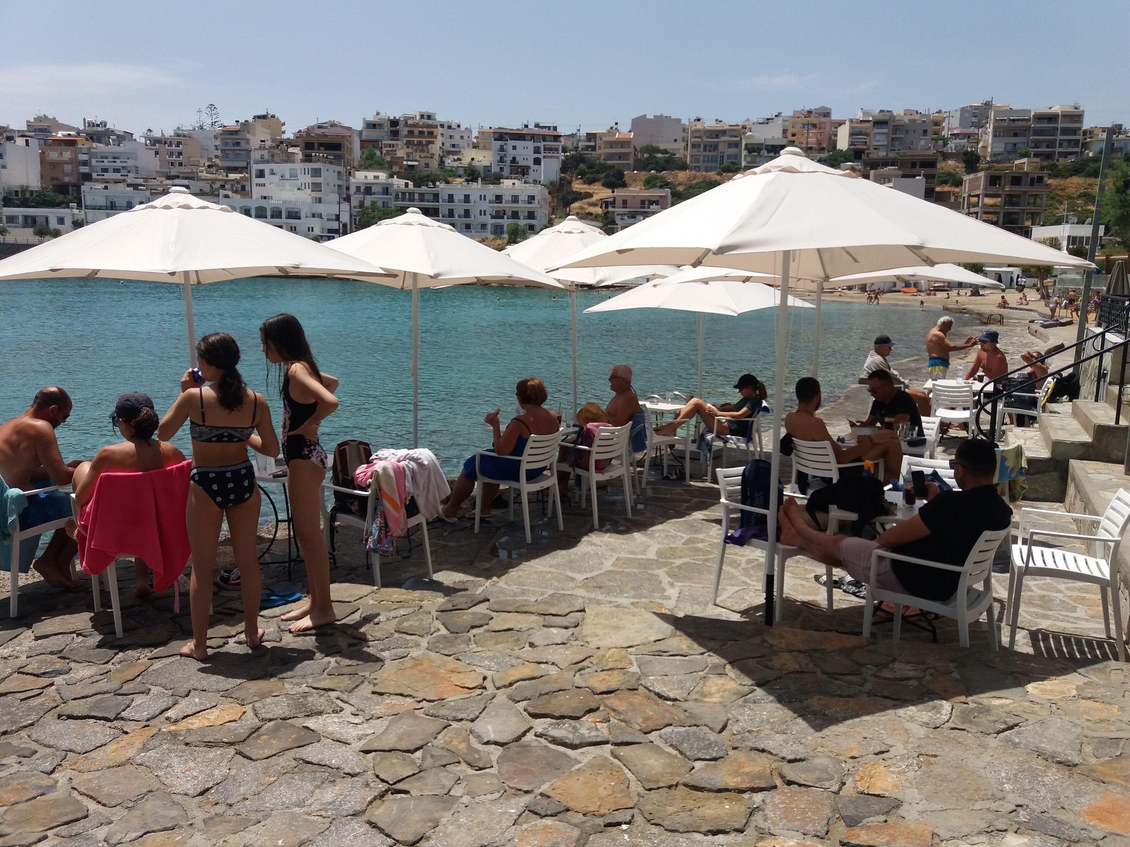Um terraço costeiro com soalho de pedra, guarda-sóis brancos e pessoas a relaxar perto da água.