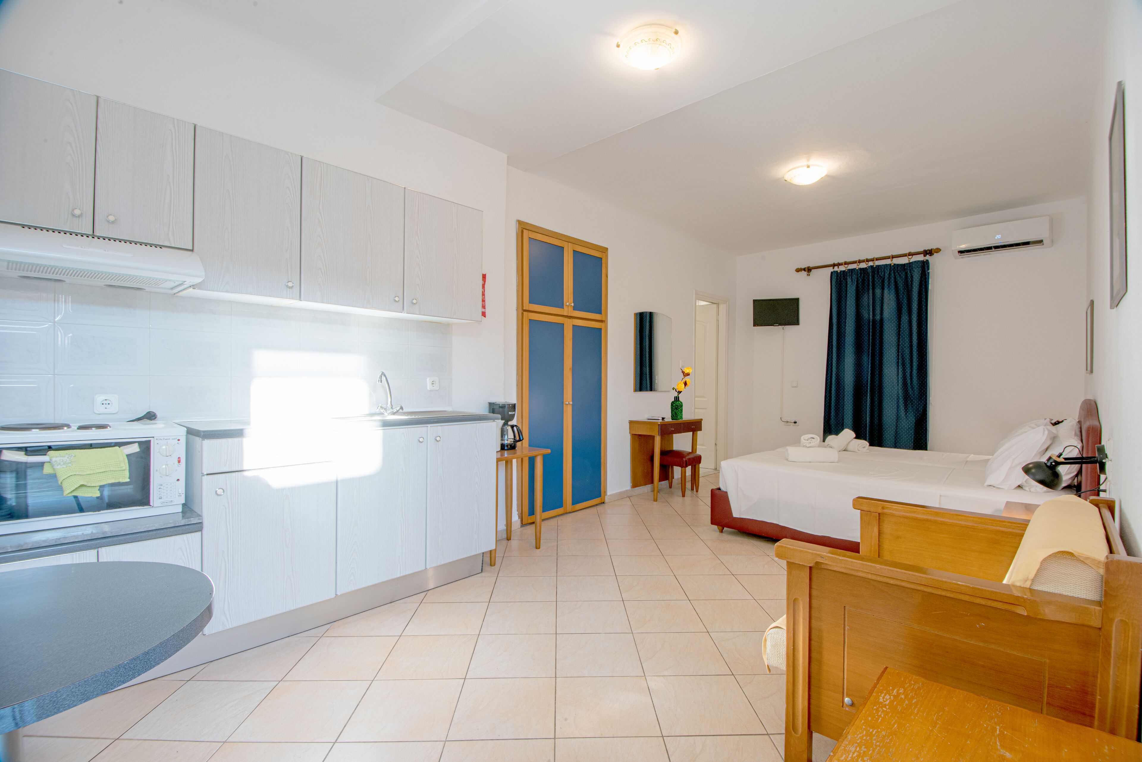 A compact hotel room with a kitchenette, a single bed, a small table, and a bedside table.
