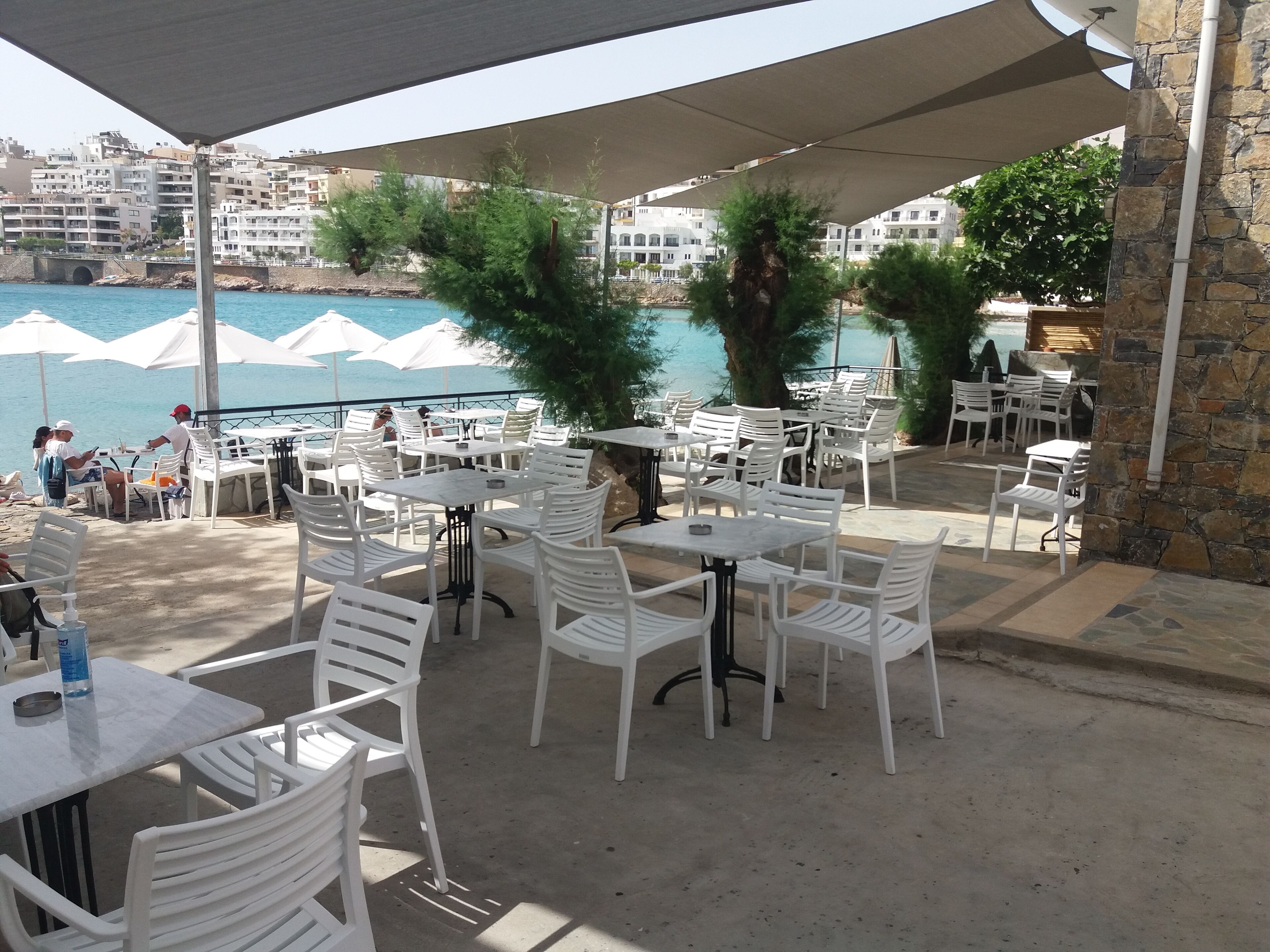 An outdoor seating area with white plastic chairs and tables, overlooking a body of water with white umbrellas.