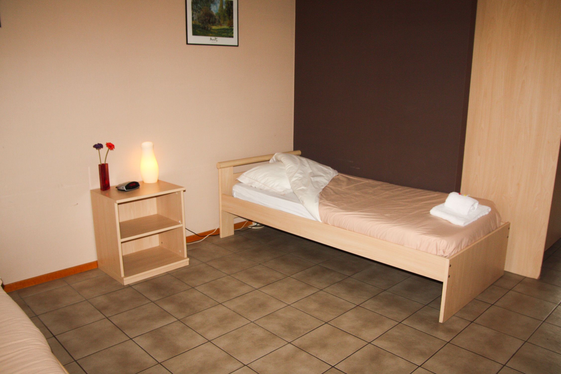 Una habitación pequeña con una cama individual, mesita de noche con lámpara y una pequeña estantería.