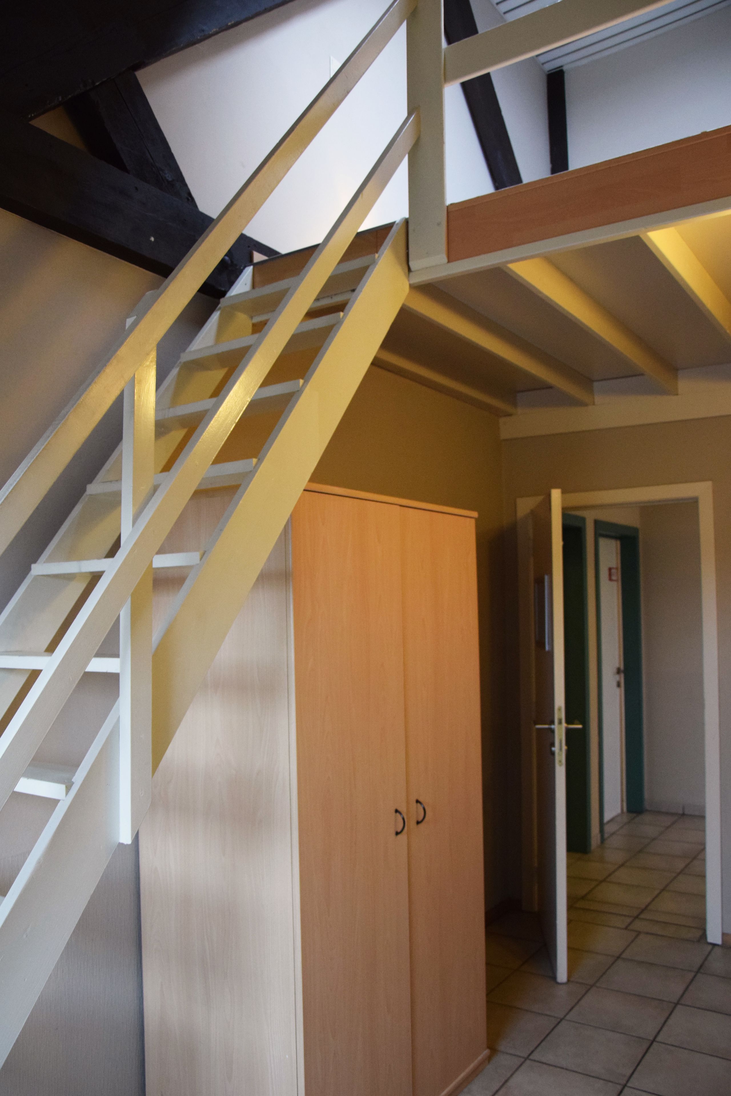 A staircase with a wooden finish and white railings, leading to a room with a wooden wardrobe and a tiled floor.