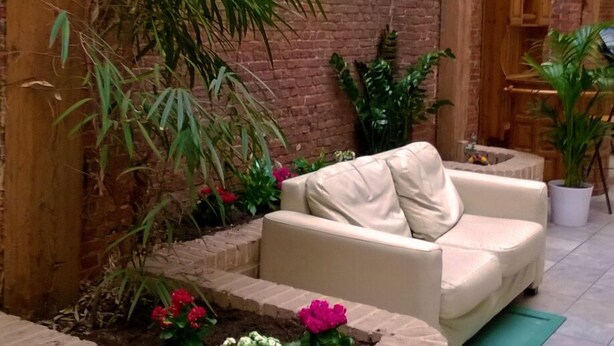 Un patio acogedor con pared de ladrillo, vigas de madera, un banco acolchado y plantas en macetas.