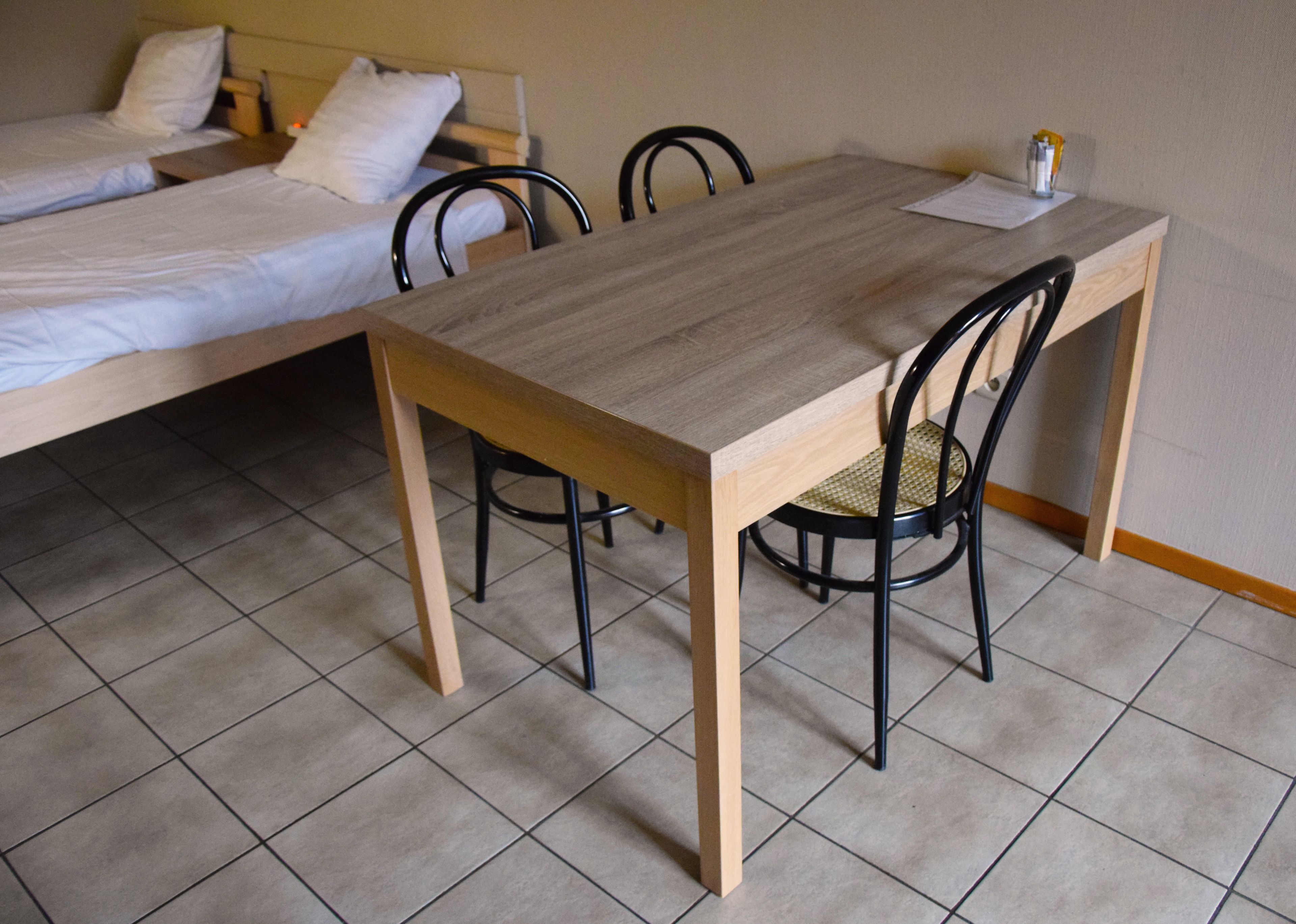 Een kamer met een houten tafel en vier stoelen, een bed met witte lakens en een betegelde vloer.
