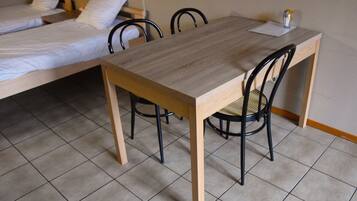Een kamer met een houten tafel en vier stoelen, een bed met witte lakens en een betegelde vloer.
