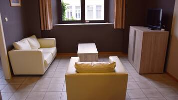 A living room with a sofa, a smaller armchair, a coffee table, a TV on a cabinet, and a window with curtains.