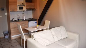Una kitchenette compacta con microondas, pileta y refrigerador pequeño. Hay un sofá blanco y una mesa de comedor con sillas en la sala.