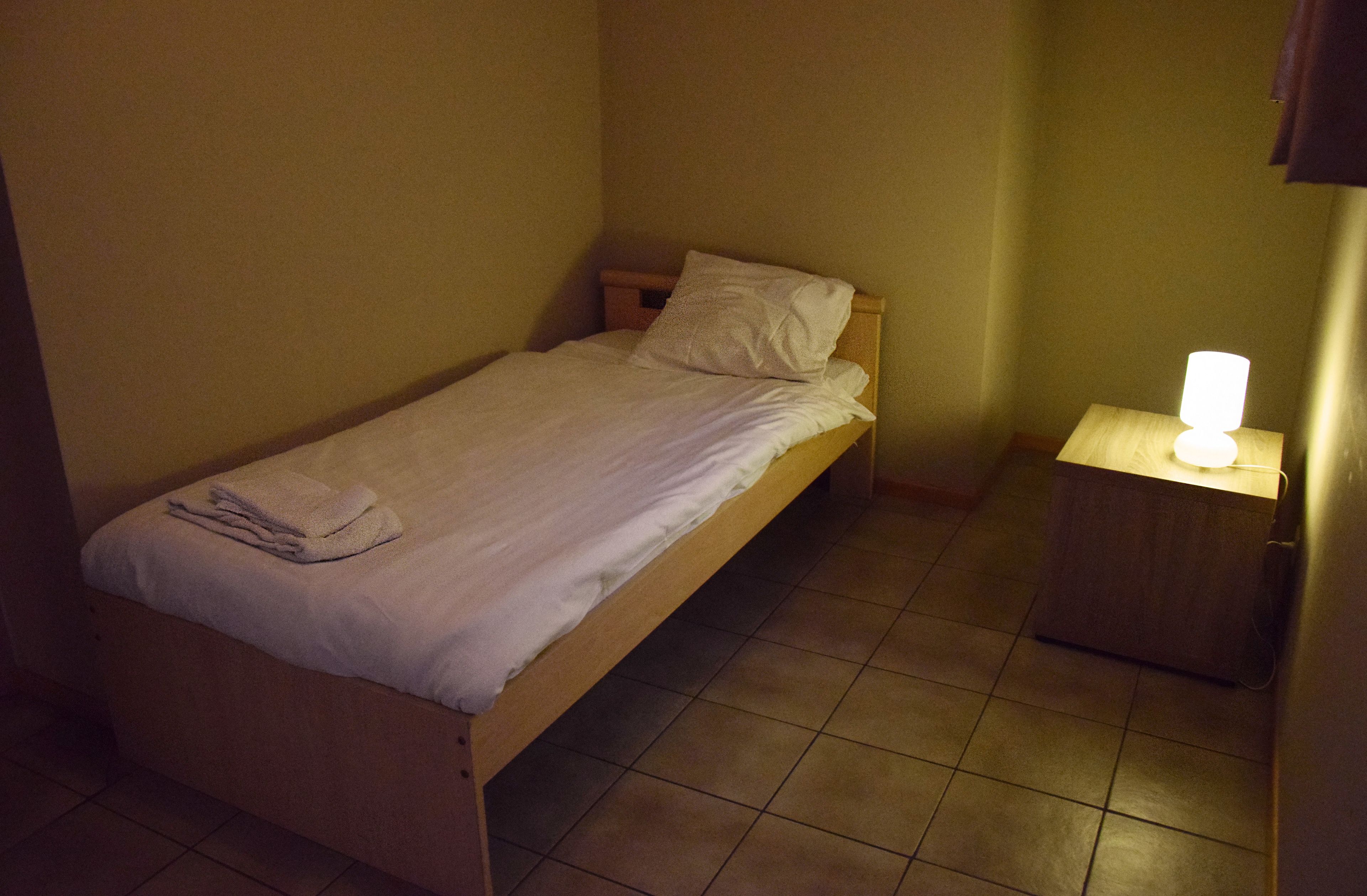 Una cama individual con sábana blanca y almohada, una toalla doblada sobre la cama y una mesita de noche con una lámpara.