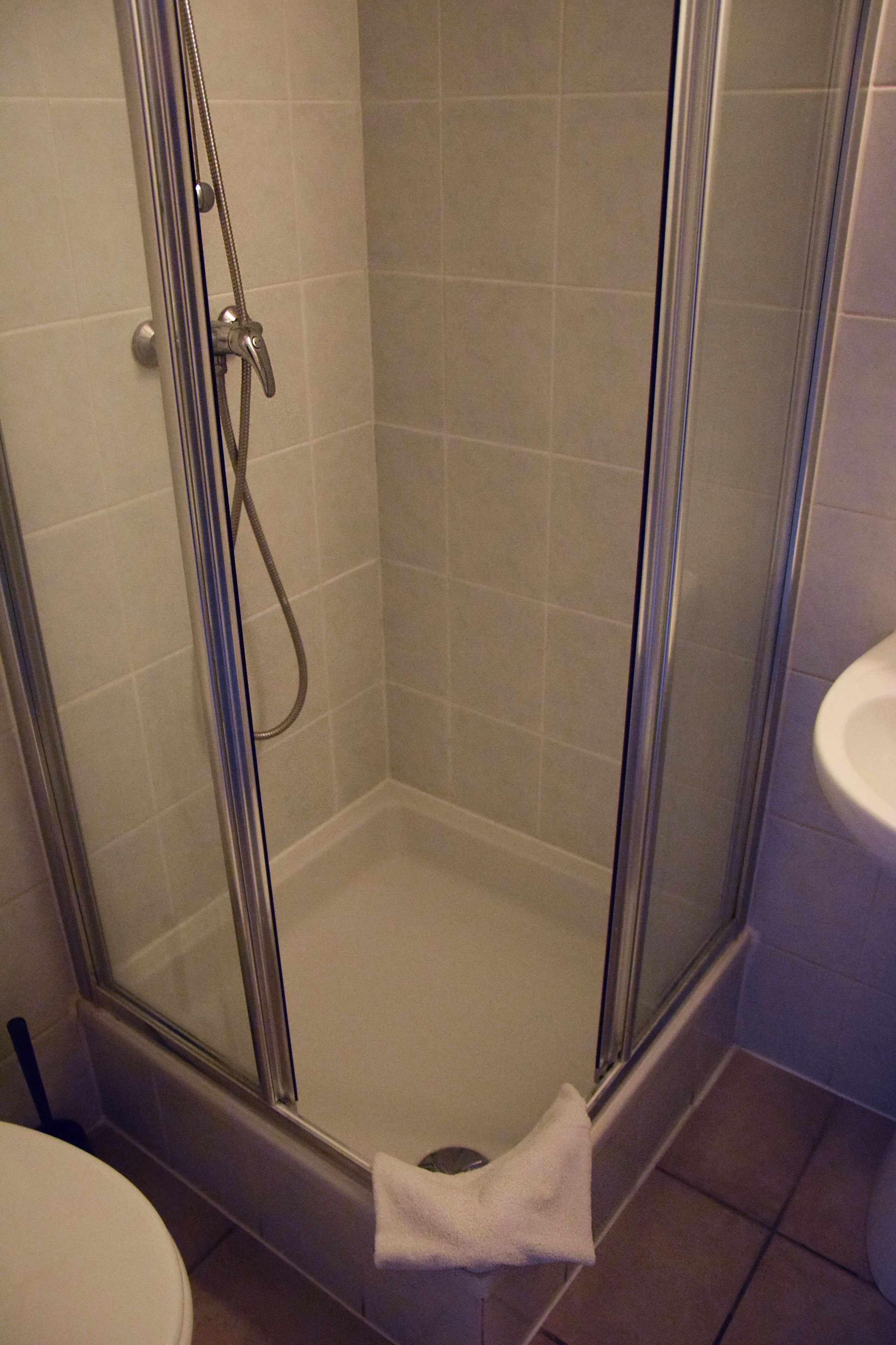 Eine Duschkabine mit Glastüren, ein weißes Handtuch auf dem Boden, ein weißes Waschbecken und geflieste Wände.
