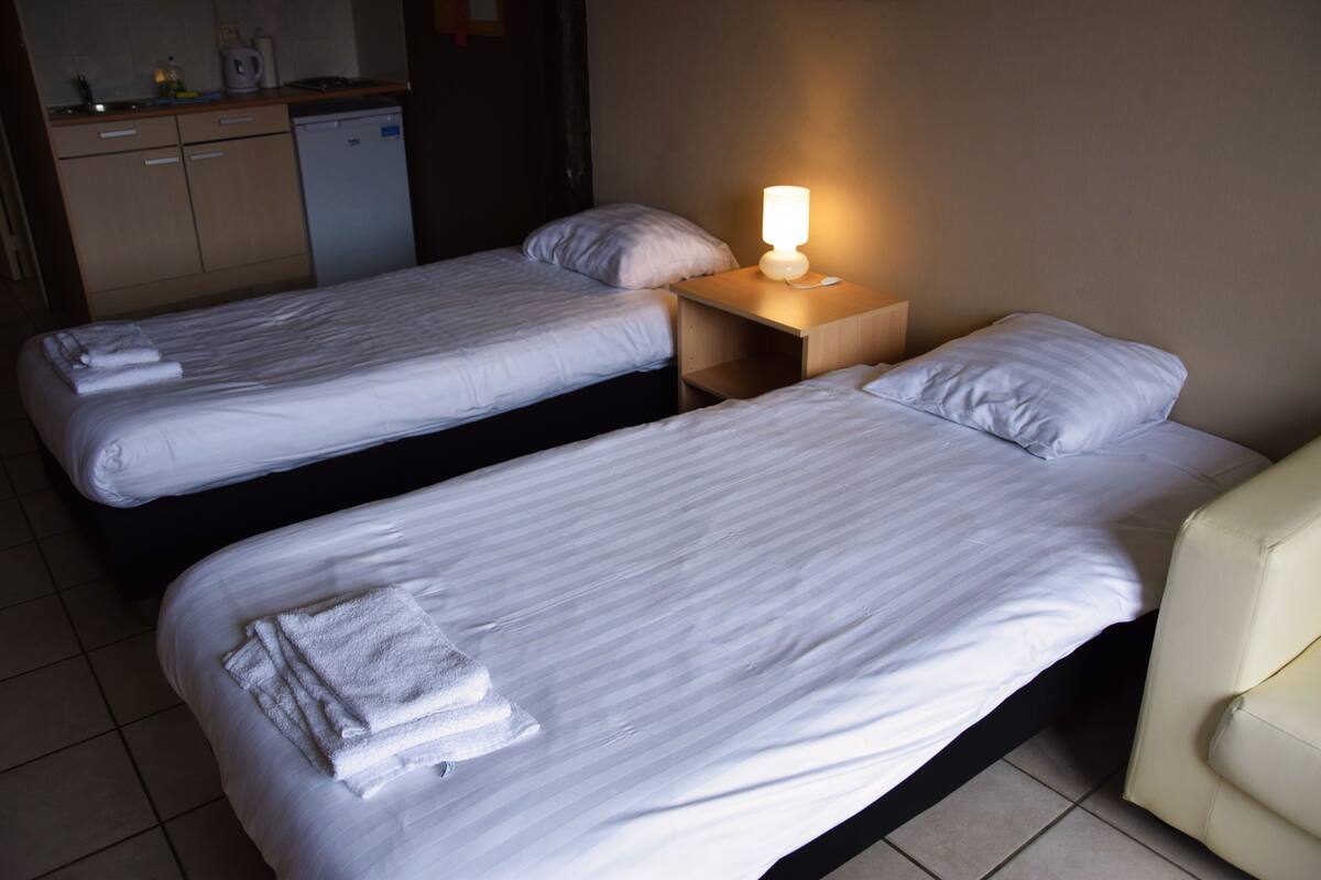 Two single beds with white linens and towels, a bedside table with a lamp, and a small kitchenette in the background.