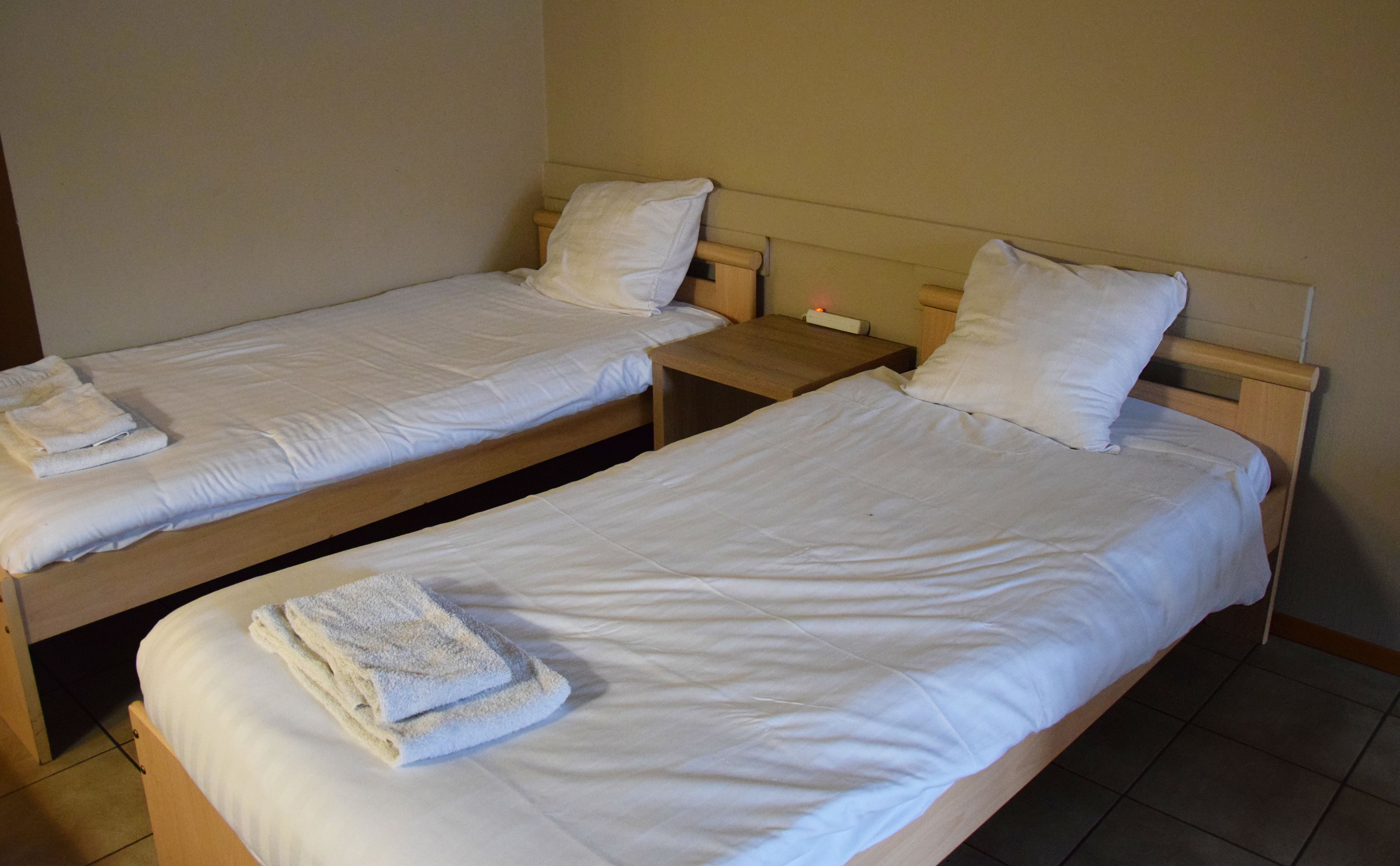 Dos camas individuales con sábanas y almohadas blancas, cabeceros de madera y una pequeña mesita de noche con una lámpara entre ellas.