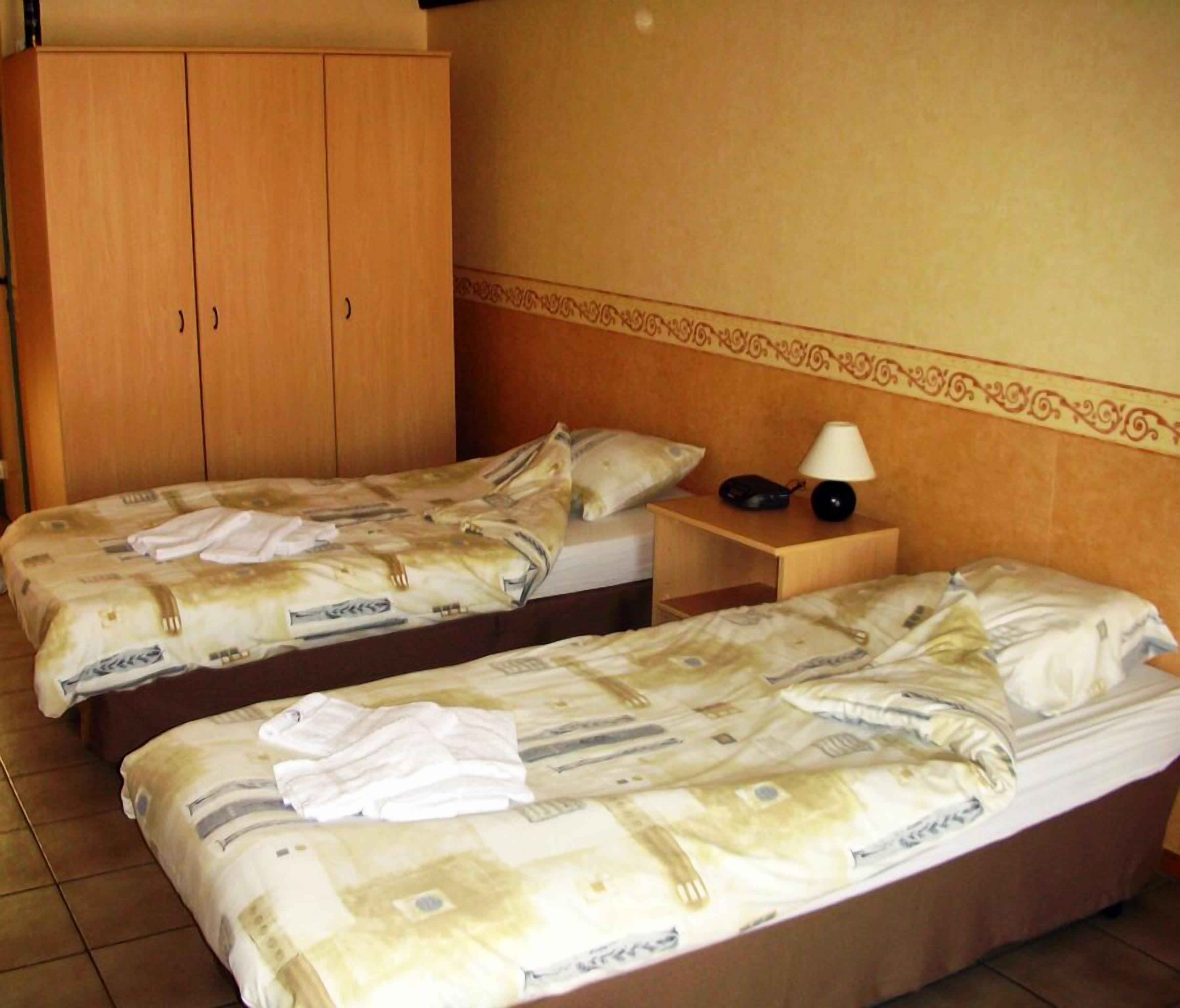A hotel room with two single beds, a wooden wardrobe, a nightstand with a lamp, and a decorative wall border.