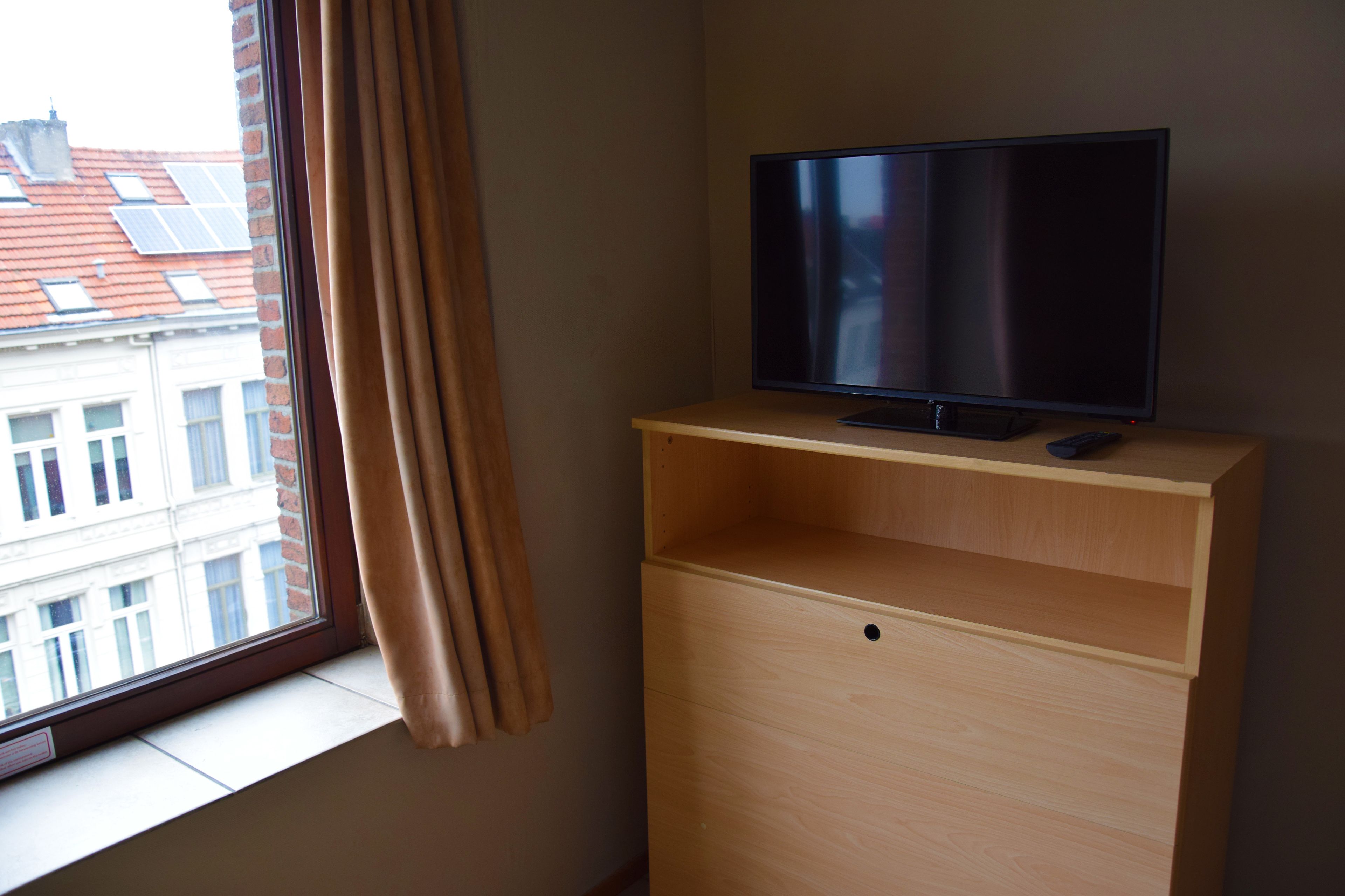 Une pièce avec un téléviseur posé sur un meuble en bois, une fenêtre avec des rideaux et une vue sur les bâtiments à l’extérieur.