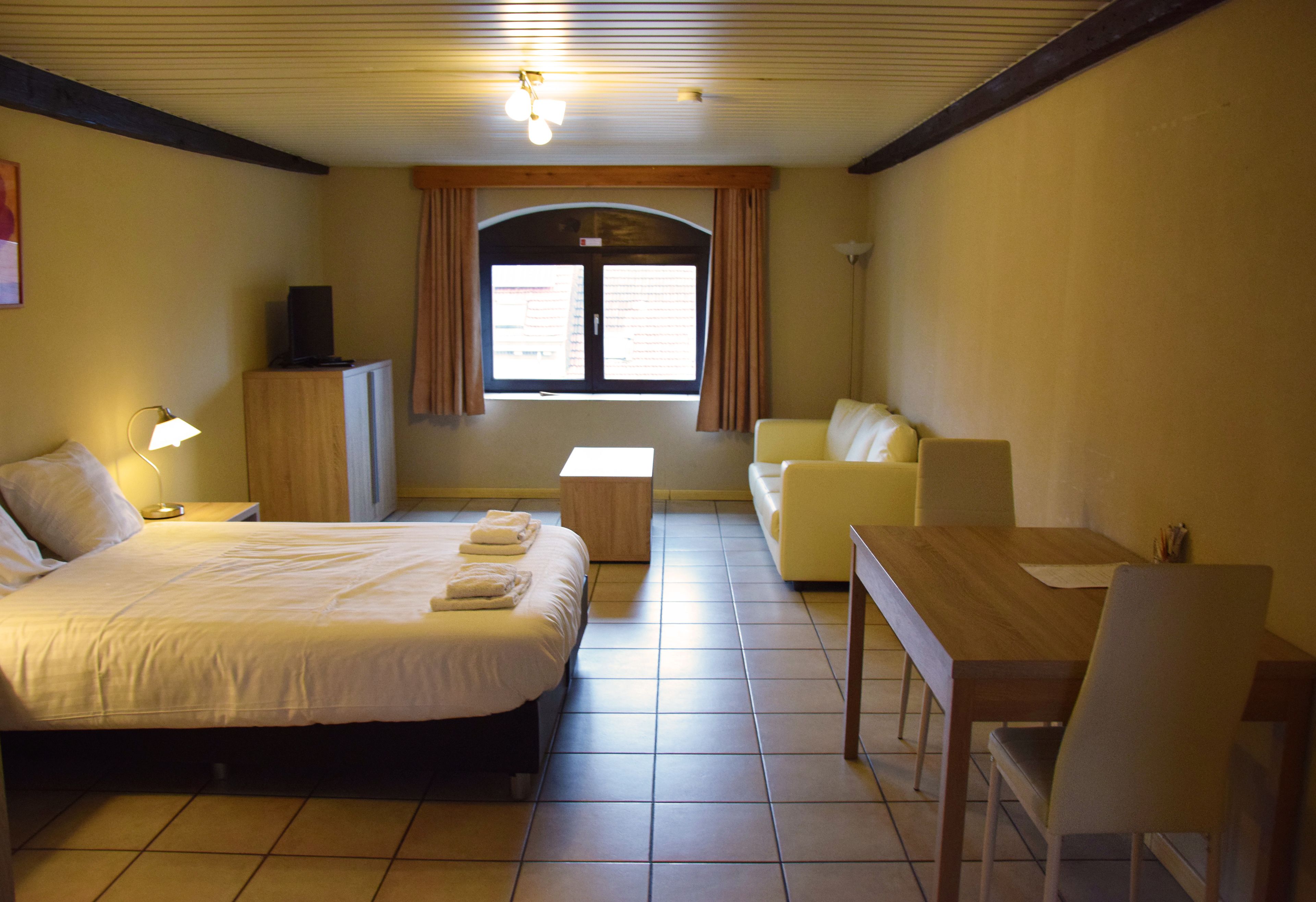Habitación de hotel con cama, escritorio, silla, sofá, televisor en un mueble y ventana con cortinas.
