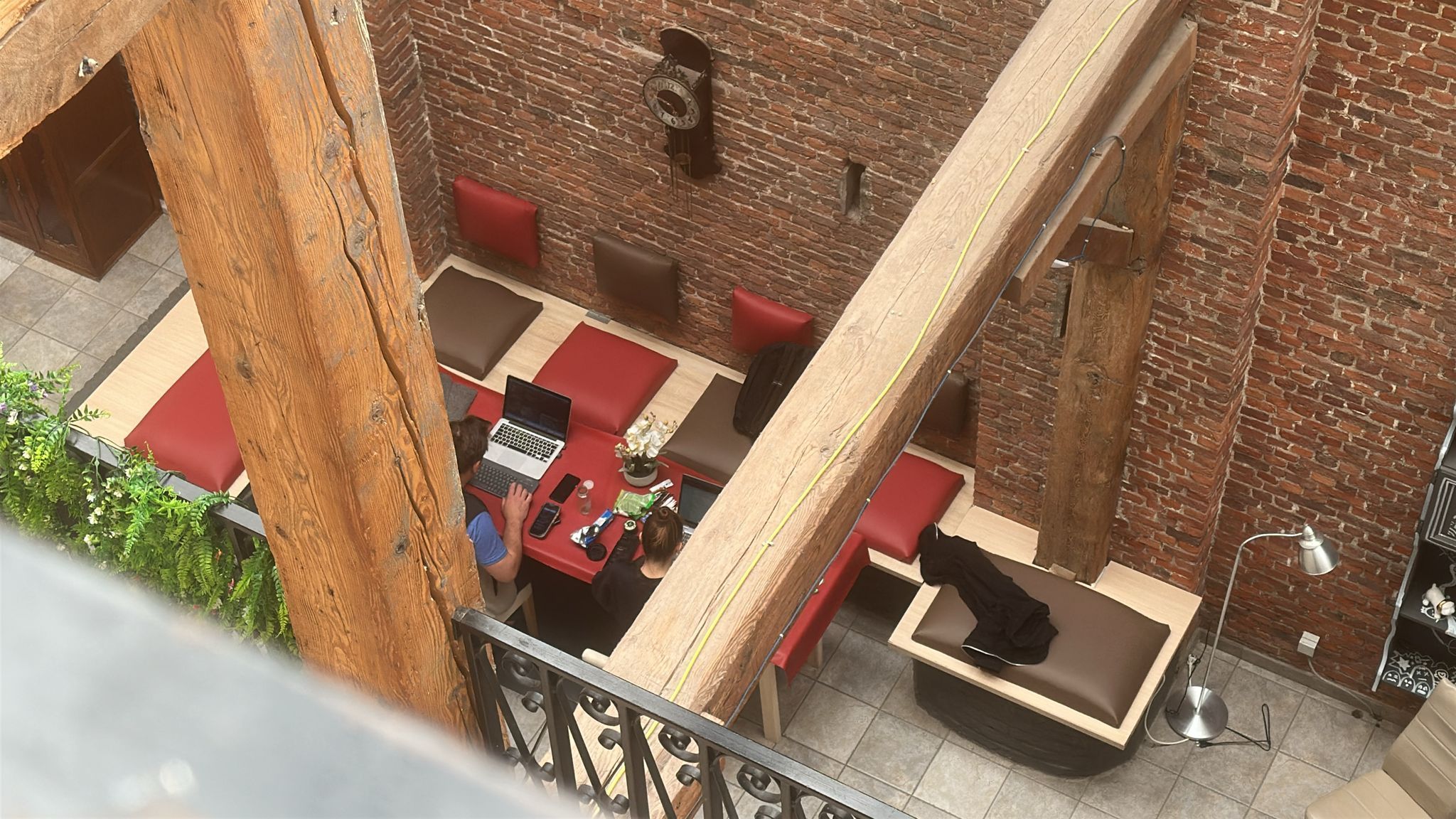 A brick-walled interior with a wooden staircase, red cushioned seating, and a table with a laptop.