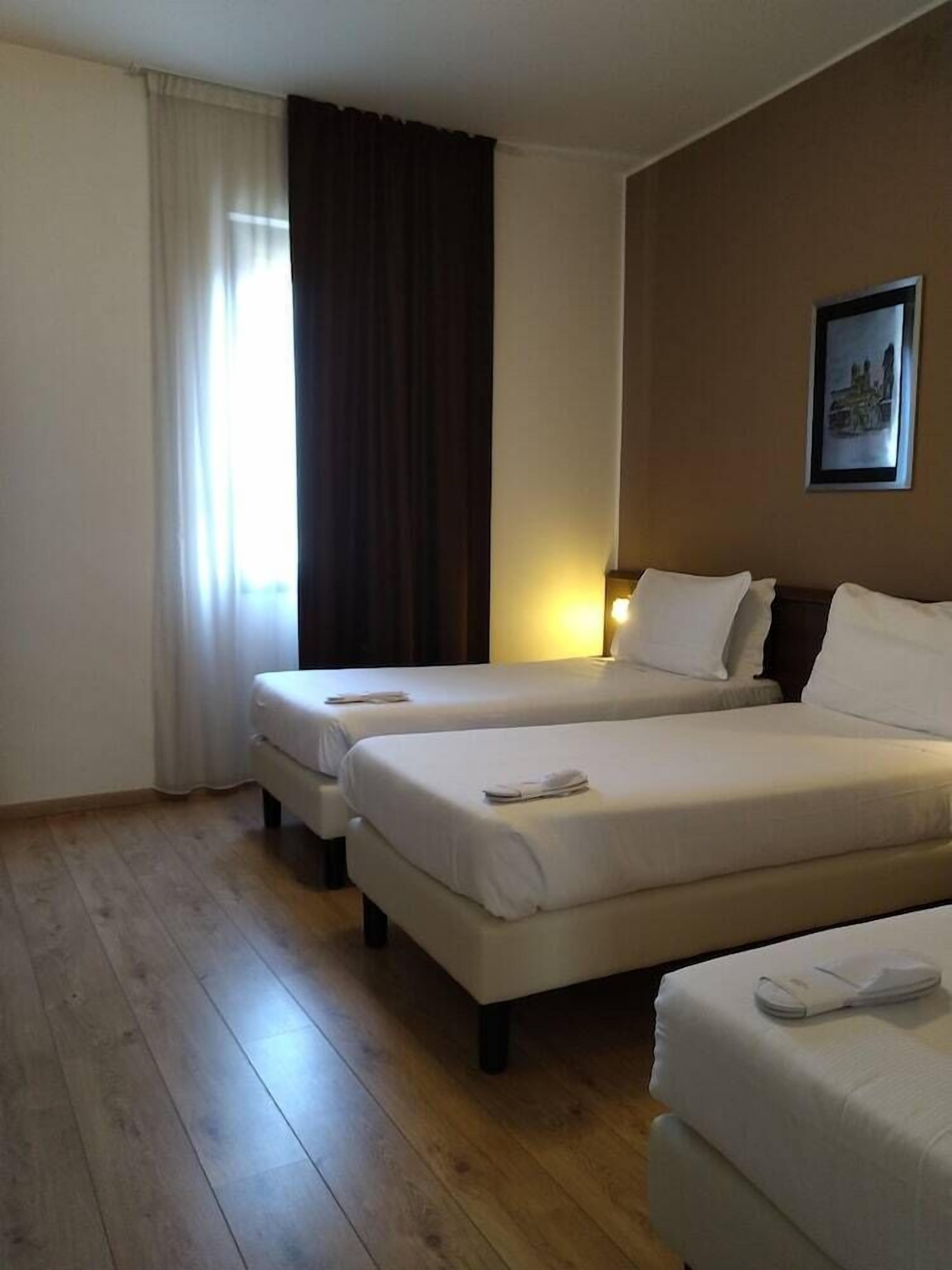 Habitación de hotel con dos camas, un ventanal con cortinas, una lámpara de pared y un cuadro enmarcado.