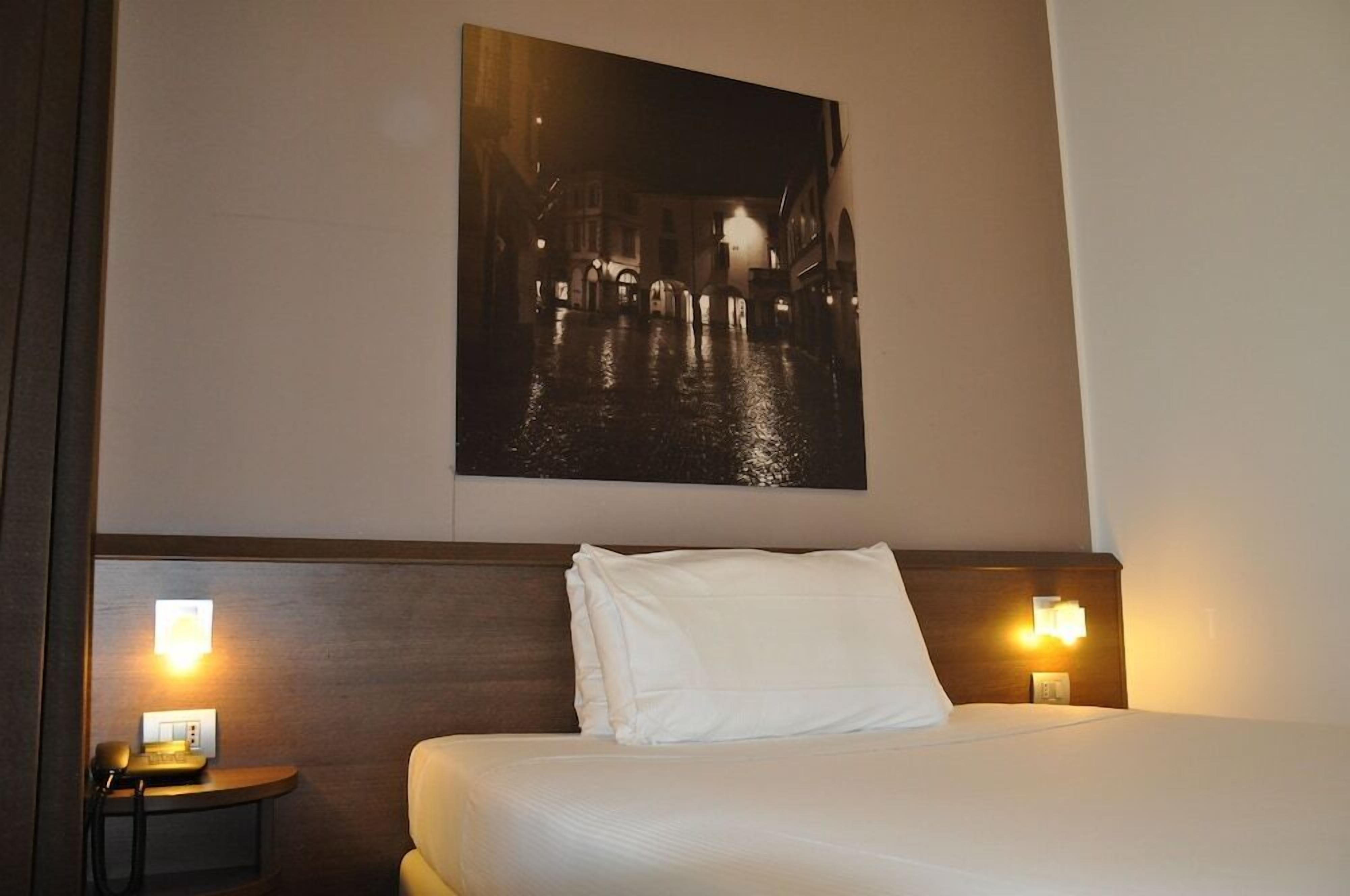 Ein Hotelzimmer mit einem Bett, zwei Nachttischlampen, einem Nachttisch und einem an der Wand hängenden Stadtbild.