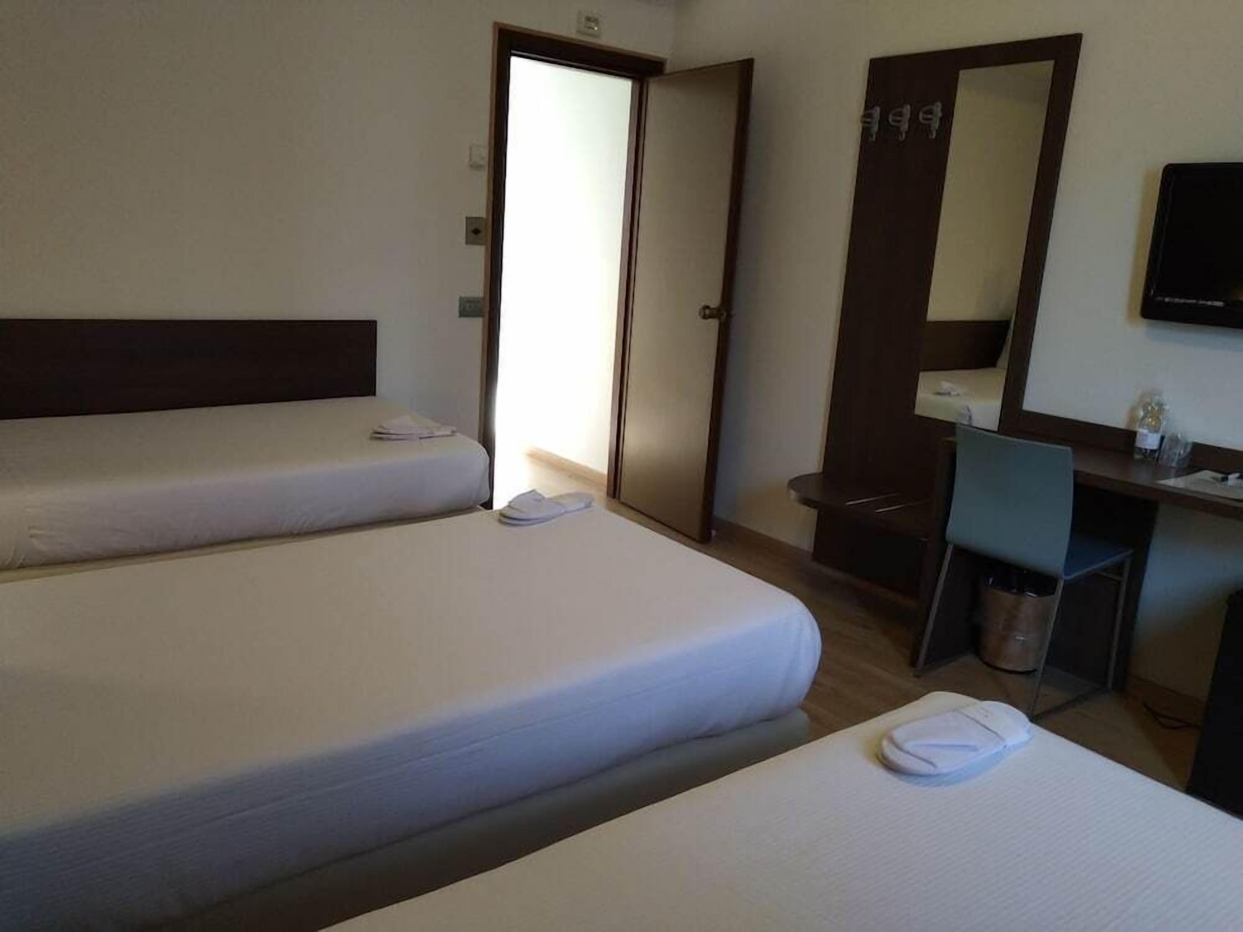 Une chambre d’hôtel avec deux lits simples, un bureau avec une chaise, un miroir et une petite poubelle.
